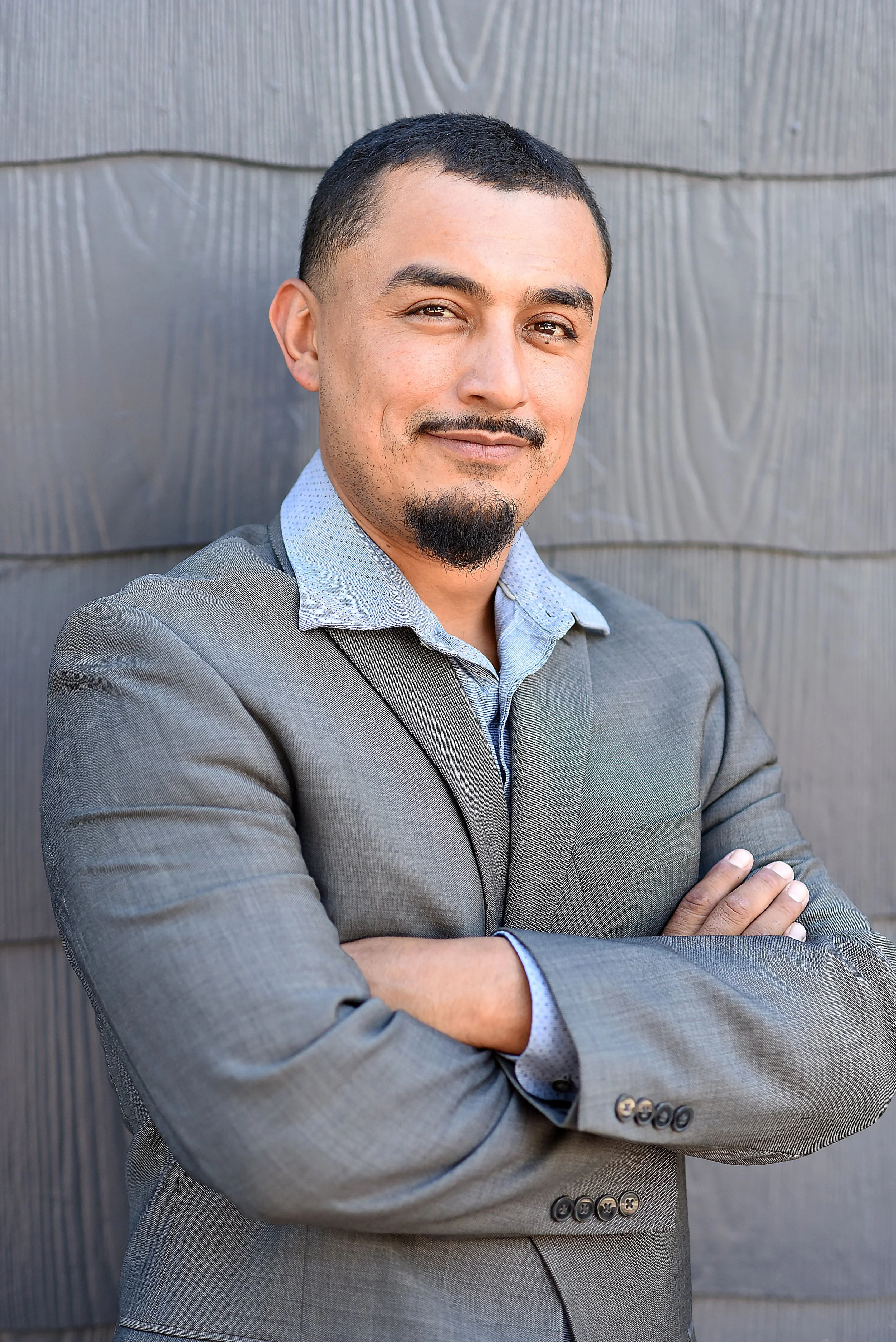 A man with short dark hair, a goatee, and light brown skin, wearing a light gray suit and blue shirt, standing against a gray textured wall with arms crossed.