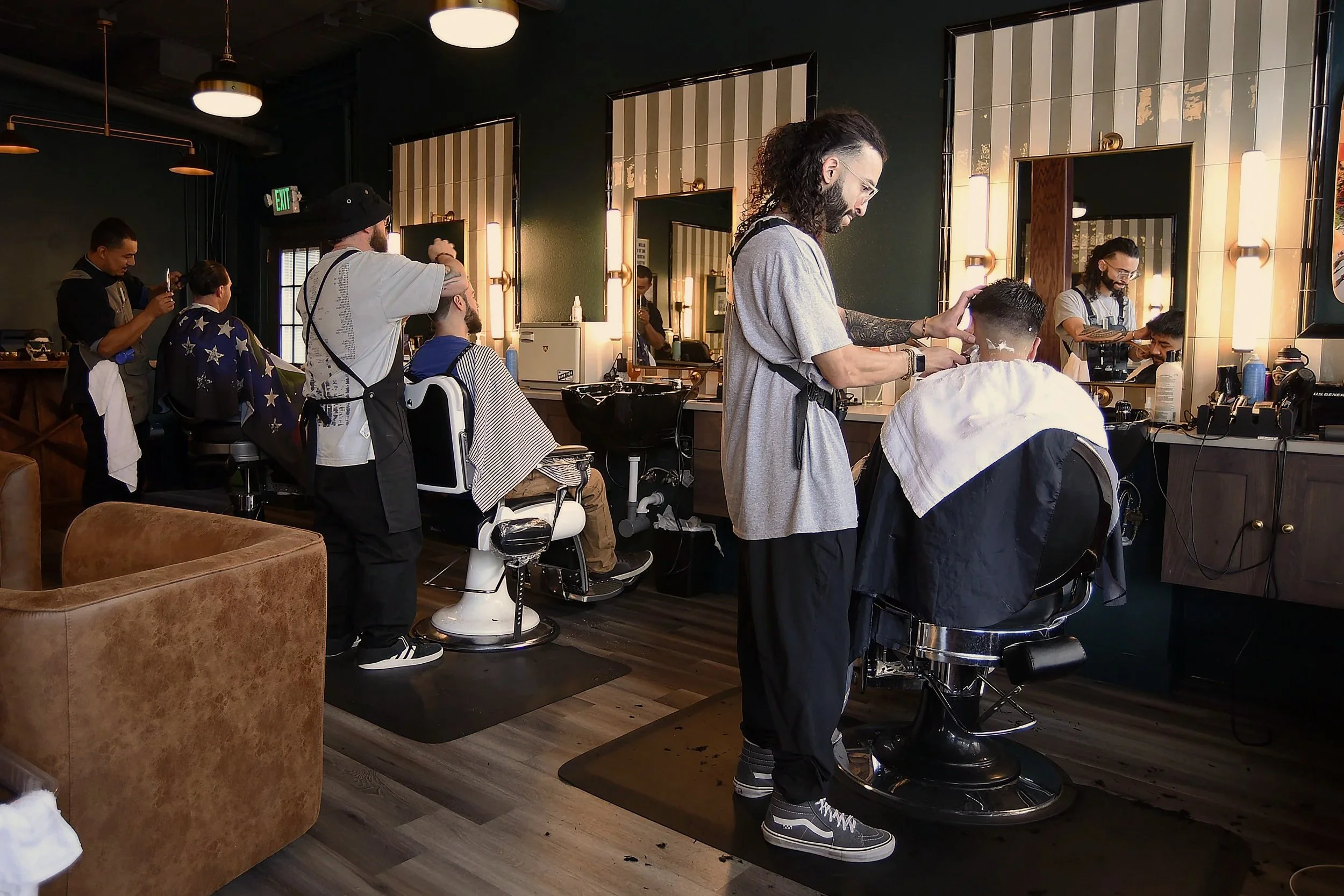 Interior of a modern barbershop with multiple hair stylists cutting men's hair, mirrors on the wall, and salon chairs, with one client actively getting a haircut.