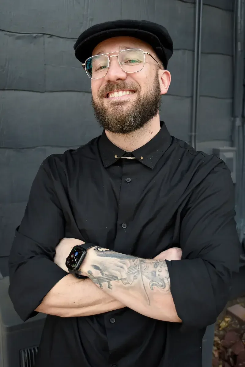A smiling man with glasses, a beard, and a black hat, wearing a black button-up shirt and a smartwatch, stands with arms crossed in front of a dark gray wall.