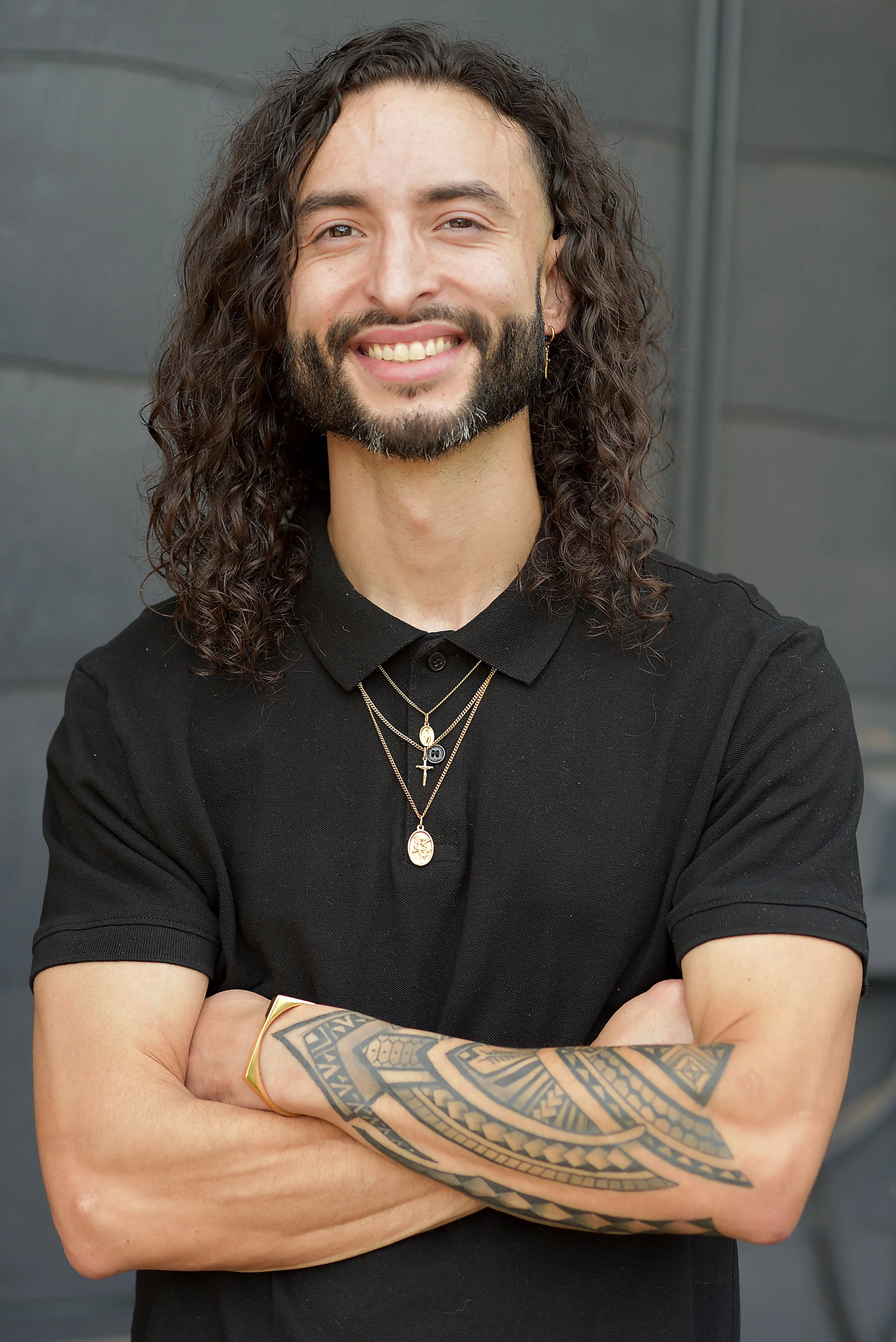 A man with long, curly dark hair and a beard, wearing a black shirt and layered gold necklaces, is smiling with arms crossed in front of a gray background. He has tattoos on his right arm and wears a gold bracelet and earrings.