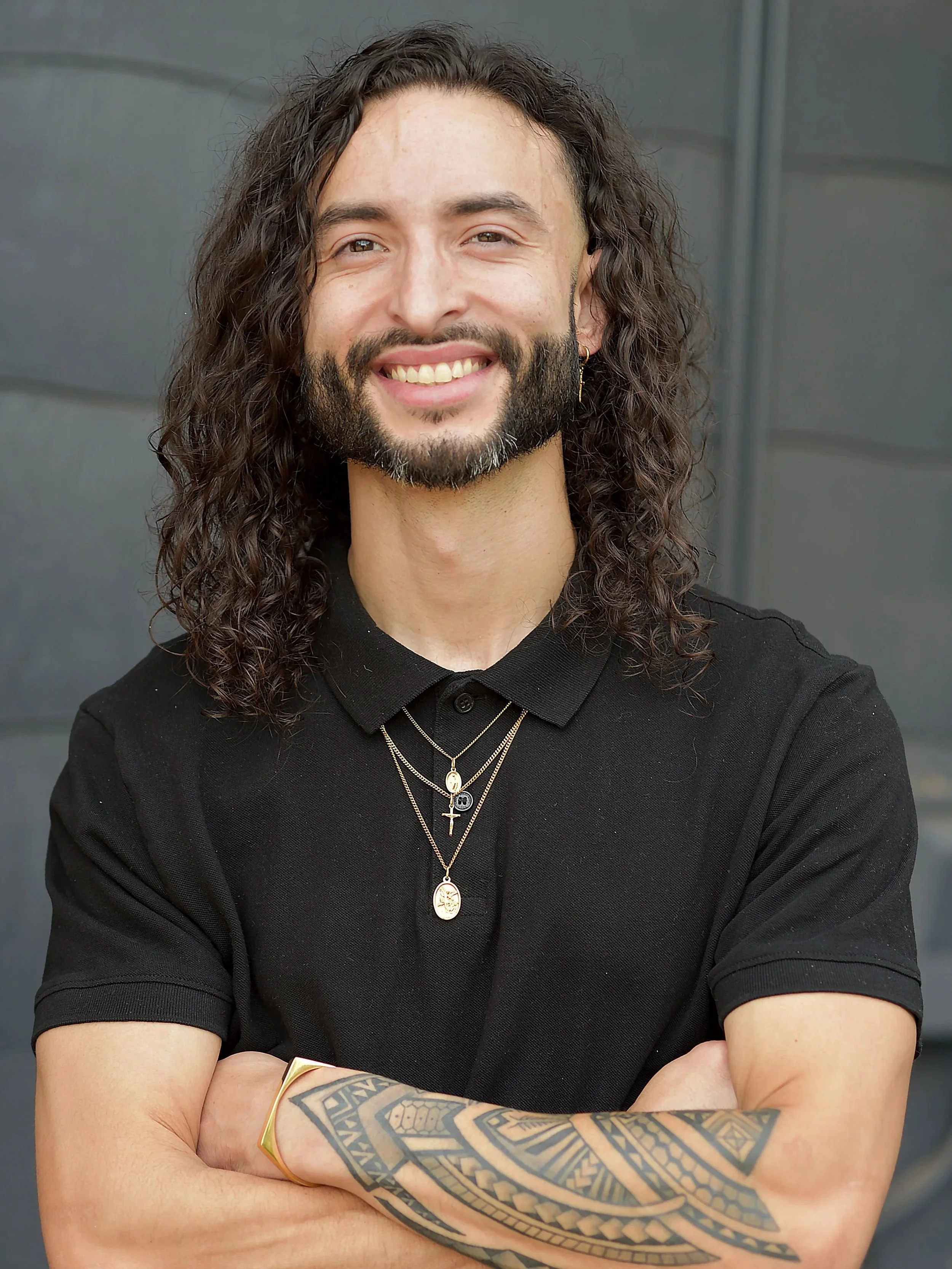A smiling man with long, curly dark hair, a beard, wearing a black polo shirt, layered necklaces, a tattooed arm, and a gold bracelet, standing against a dark background.