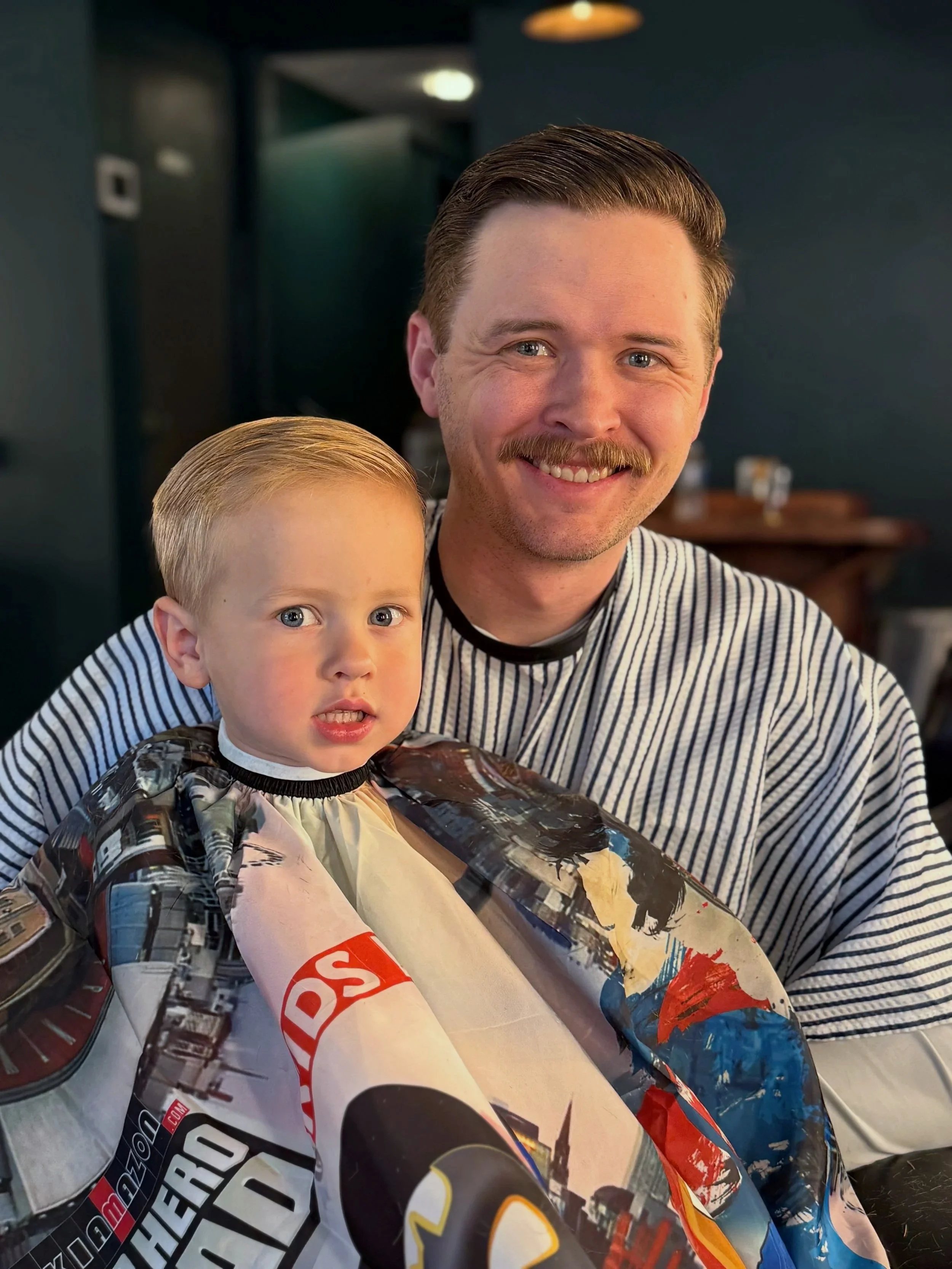 A man with a mustache and blue eyes smiling, sitting with a young boy at a barbershop. The boy has blond hair and is wearing a barber's cutting cape with super hero graphics. They are indoors with dark walls and warm lighting.
