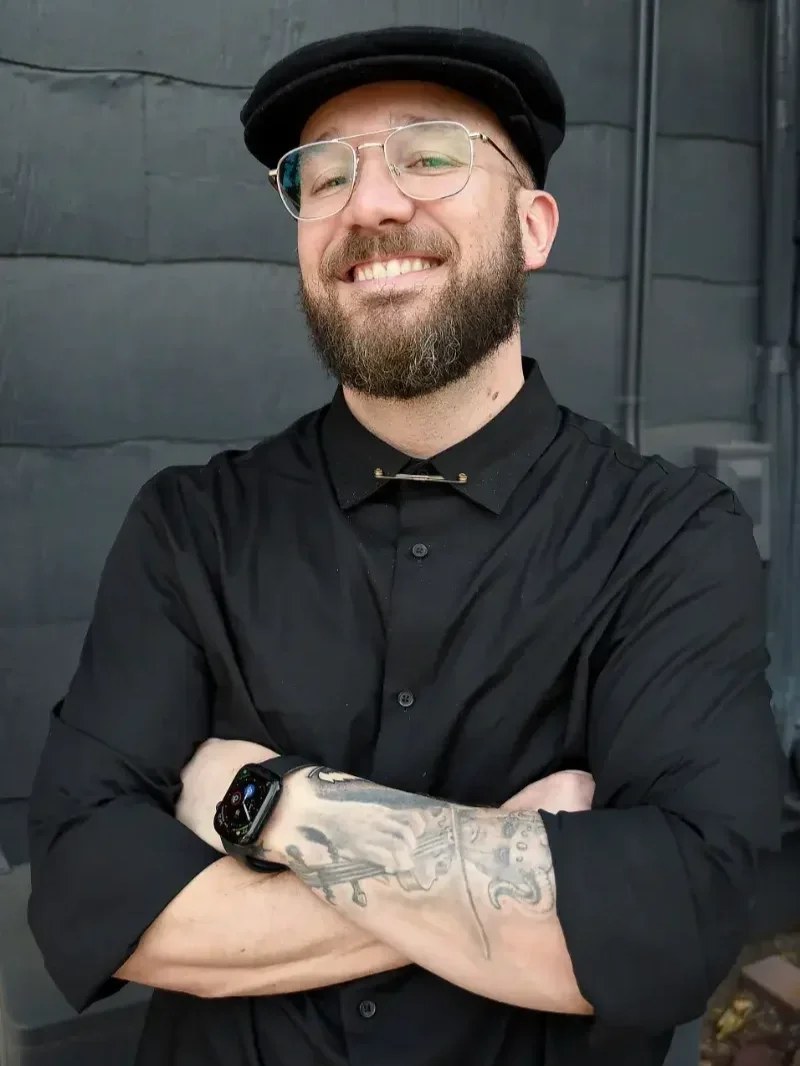 A smiling man with a beard and glasses, wearing a black shirt with rolled-up sleeves, a black cap, and a smartwatch, standing in front of a dark wall.