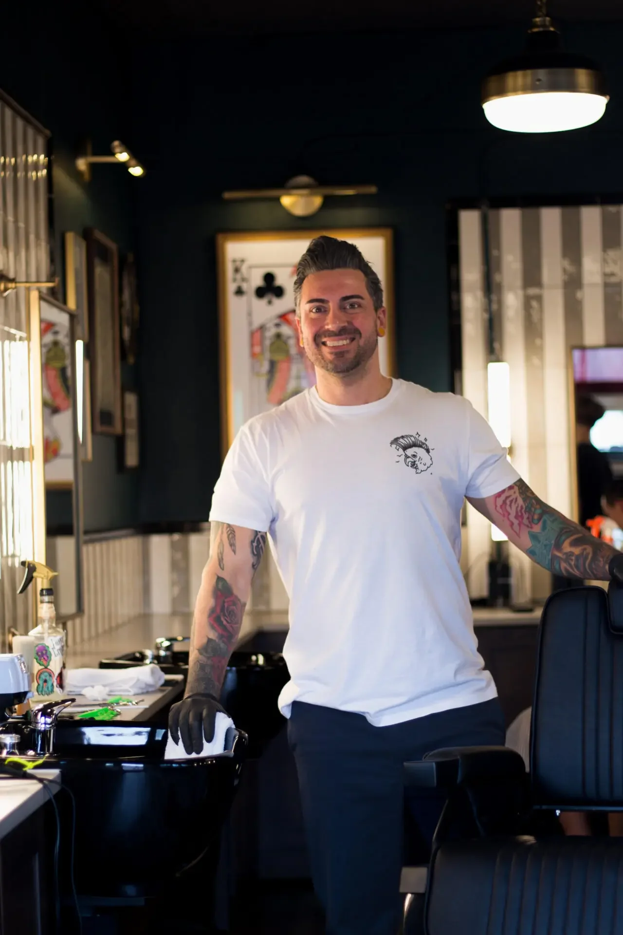 A smiling man with dark hair, tattoos on his arms, wearing a white t-shirt with a small graphic of a lion, standing in a hair salon or barbershop surrounded by tools and equipment.