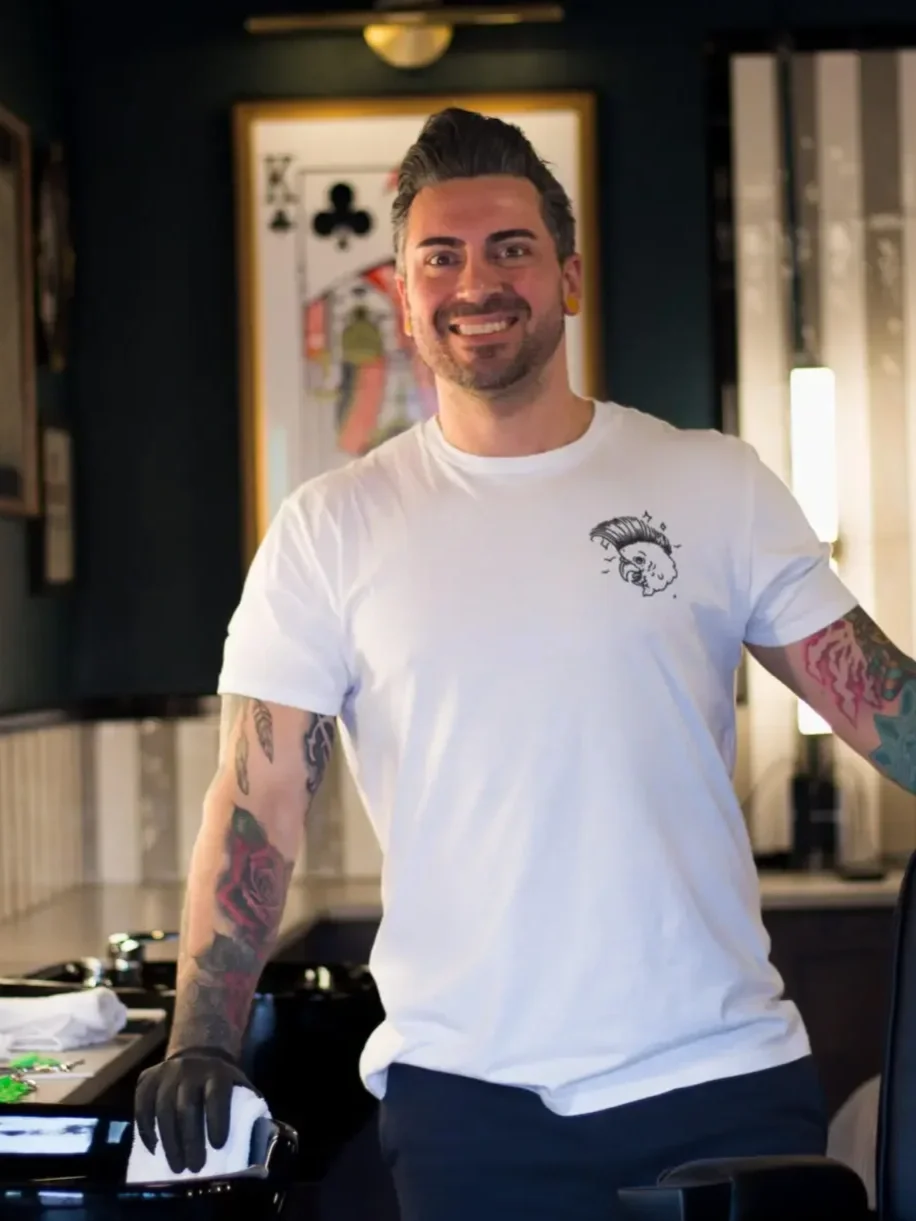 A man with dark hair styled back, smiling, wearing a white T-shirt with a tattooed arm, standing inside a barbershop with a framed poster in the background.