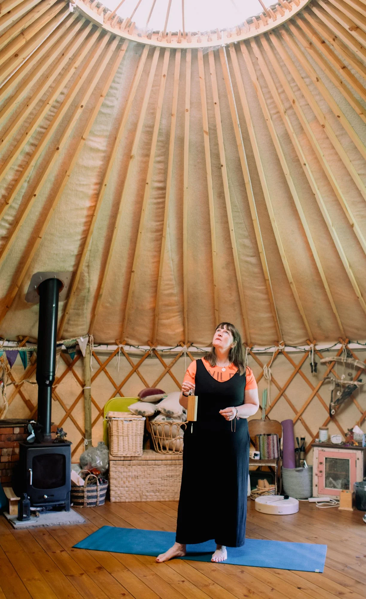 A woman standing on a blue yoga mat inside a yurt with a wooden floor, holding a koshi chime in one hand, looking up at the ceiling. The interior includes storage baskets, a wood burning stove, and books.