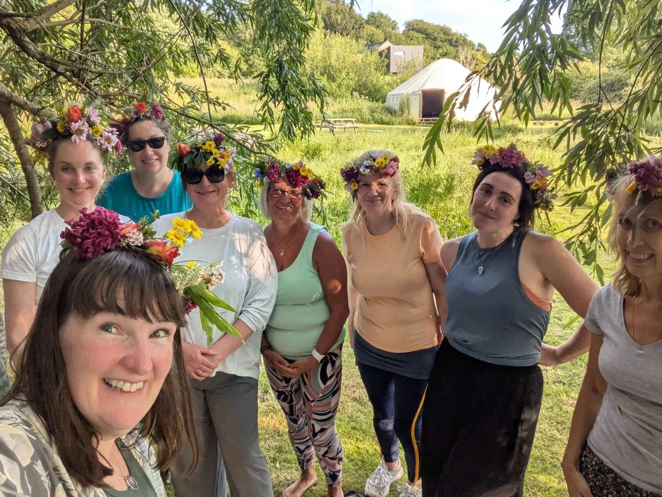 A group of women wearing floral crowns made of colorful flowers pose together outdoors in a grassy area under a willow tree and a yurt in the background.