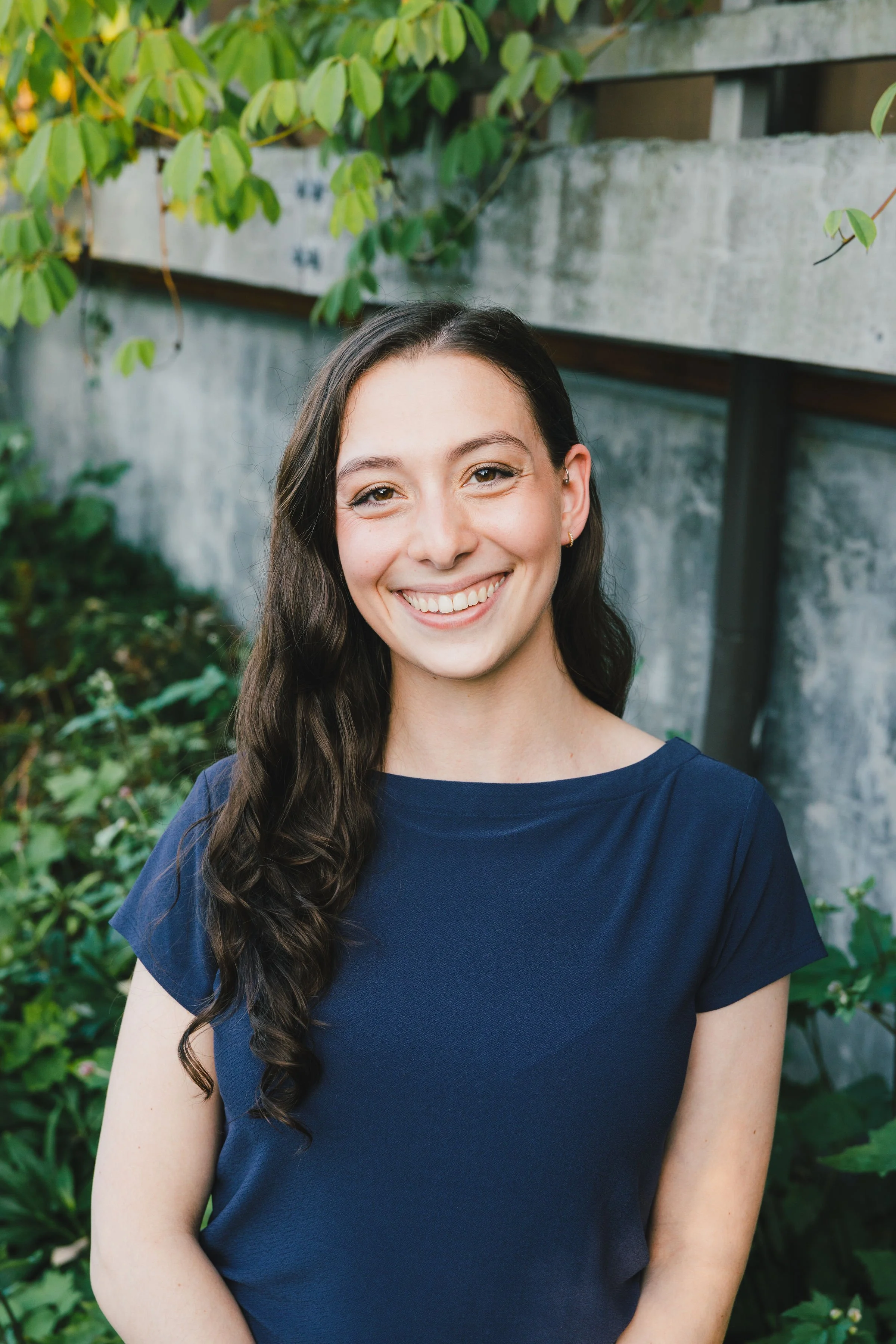 A woman with long dark hair and a blue shirt standing outdoors near greenery and a concrete wall, smiling at the camera.