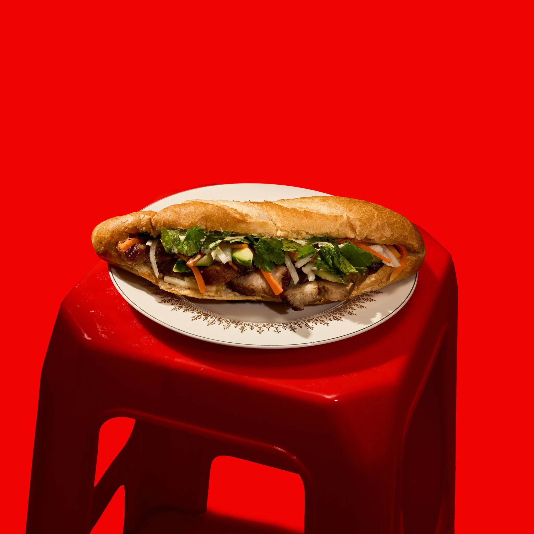 A  Vietnamese Banh Mi Sandwichewith lettuce, vegetables, and meat on a plate, against a bright red background. The photo was taken in Montréal, by Maison Toki for Pham's Banh Mi.