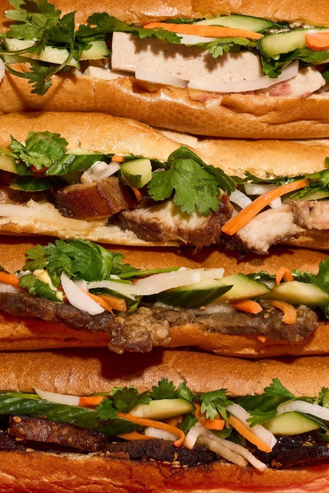 Close-up of various Vietnamese-style banh mi sandwiches with ingredients like grilled meats, fresh vegetables, cilantro, and pickled vegetables in French baguette bread.