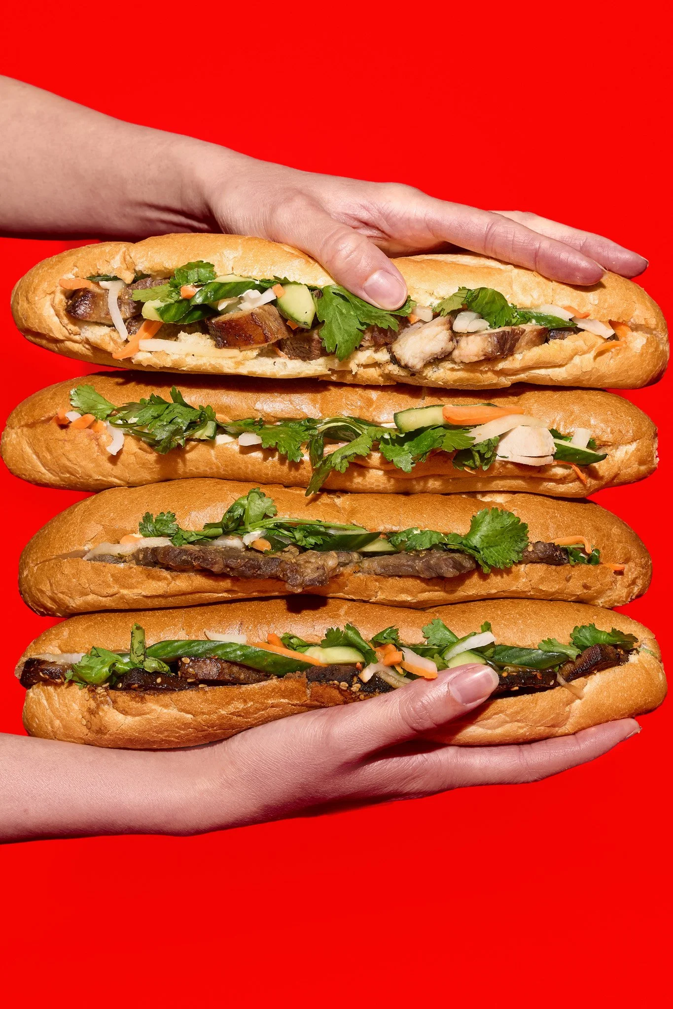 Close-up of a person holding multiple large  Bánh Mì  sandwiches with various ingredients, including vegetables and meats, over a bright red background. The photo was taken in Montréal, by Maison Toki for Pham's Banh Mi.