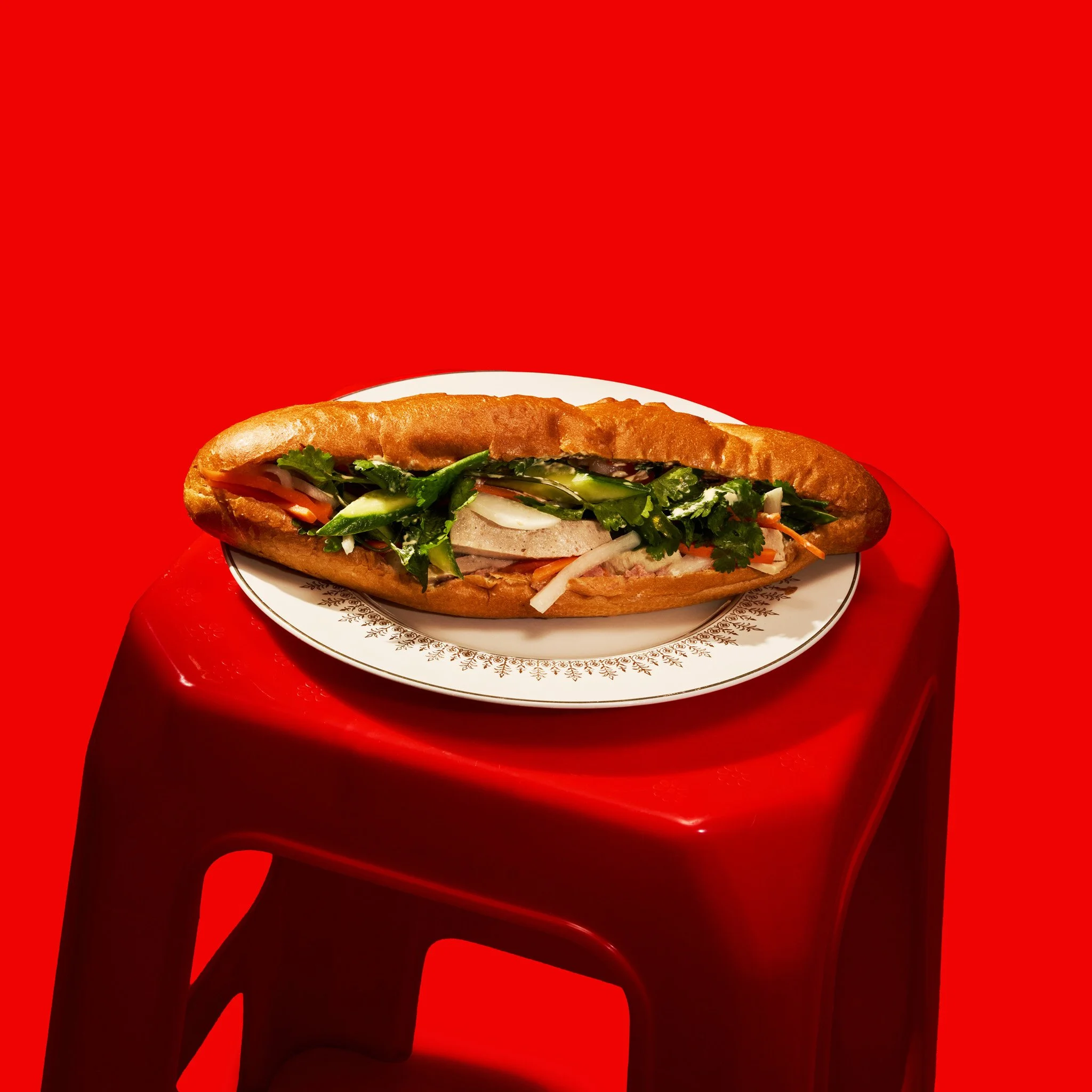 Close-up of a Vietnamese Banh Mi Sandwiche  with vegetables on a white plate, held by a person's hand against a bright red background.  The photo was taken in Montréal, by Maison Toki for Pham's Banh Mi.