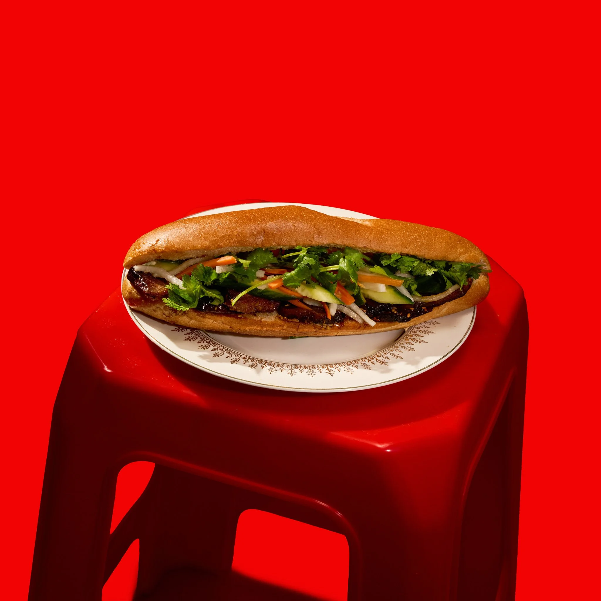 A  Vietnamese Banh Mi Sandwiche with lettuce, tomato, pickles, onions, and sliced meat or poultry, placed on a decorative plate against a red background. The photo was taken in Montréal, by Maison Toki for Pham's Banh Mi.