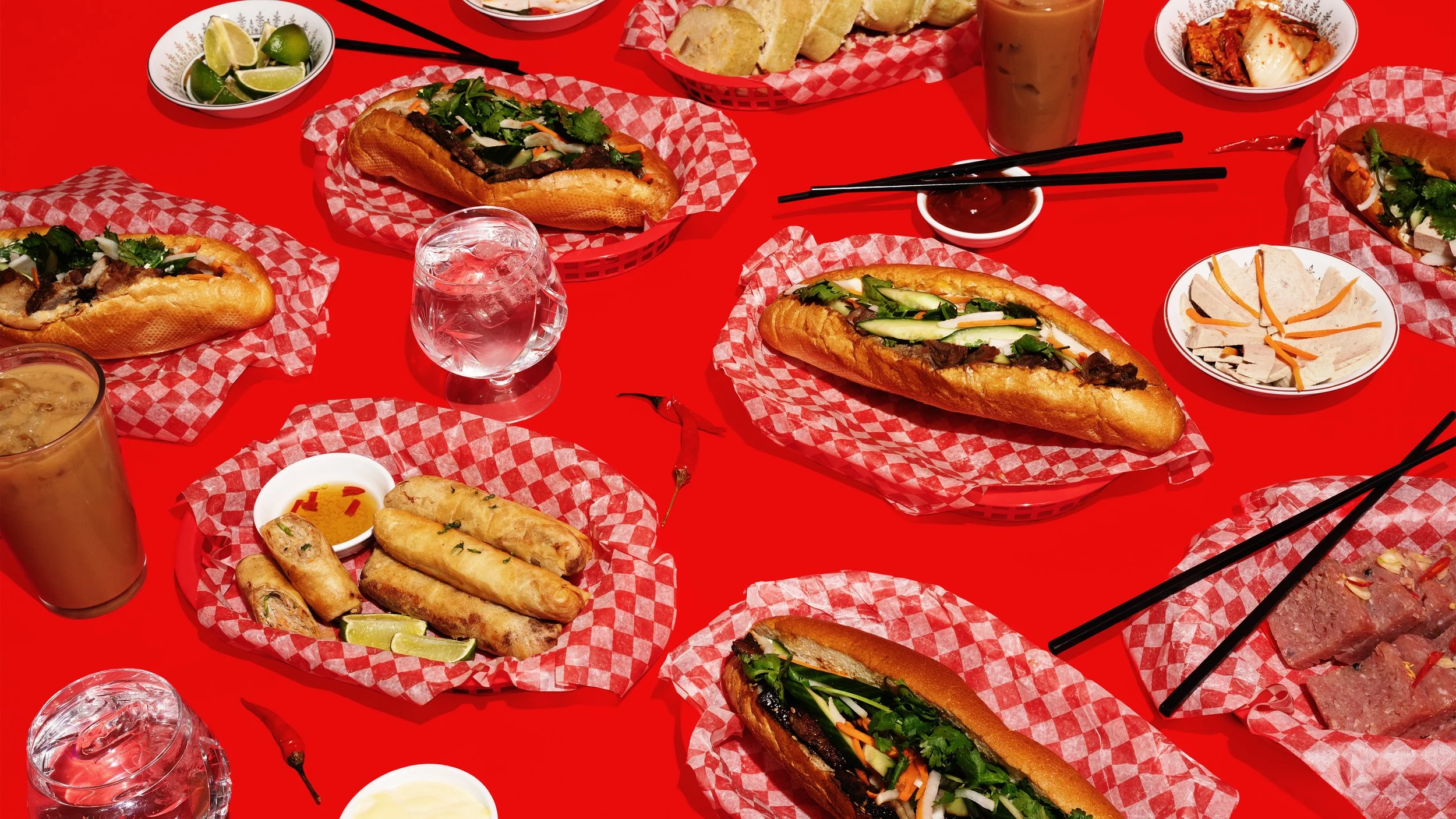 A table filled with Vietnamese dishes including multiple Banh Mi sandwiches with various fillings, small bowls of dipping sauces, lime wedges, spring rolls, side dishes, and drinks, all set on a bright red table with red checkered paper liners.