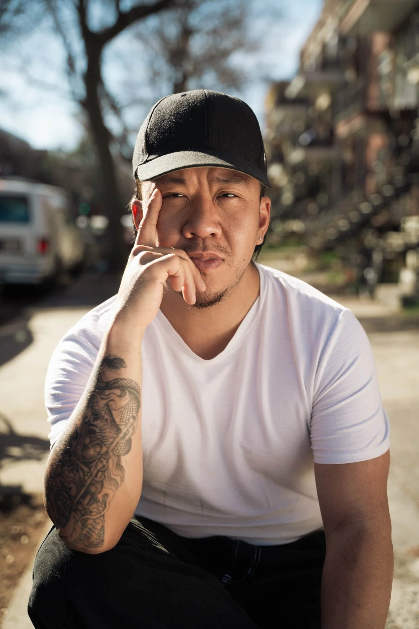 The man behind Pham's Bánh Mì sitting outdoors, wearing a black baseball cap, white t-shirt, black pants, with a tattoo on his right forearm, touching his face with his right hand, with a blurred background of trees and buildings.