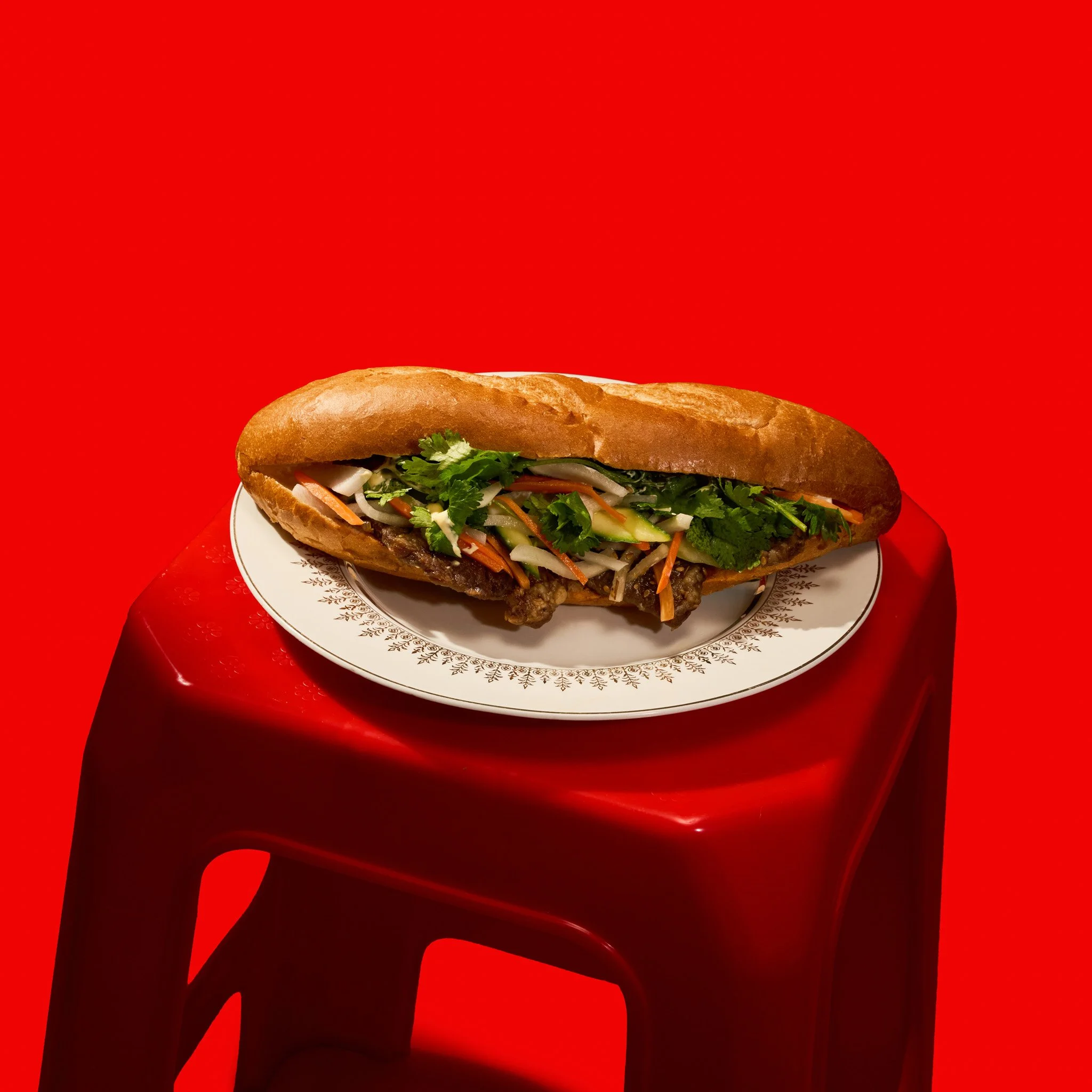 A Vietnamese Banh Mi Sandwiche with lettuce, tomato, onion, and other ingredients in a long bun, placed on a decorative plate with a floral pattern, against a red background. The photo was taken in Montréal, by Maison Toki for Pham's Banh Mi.