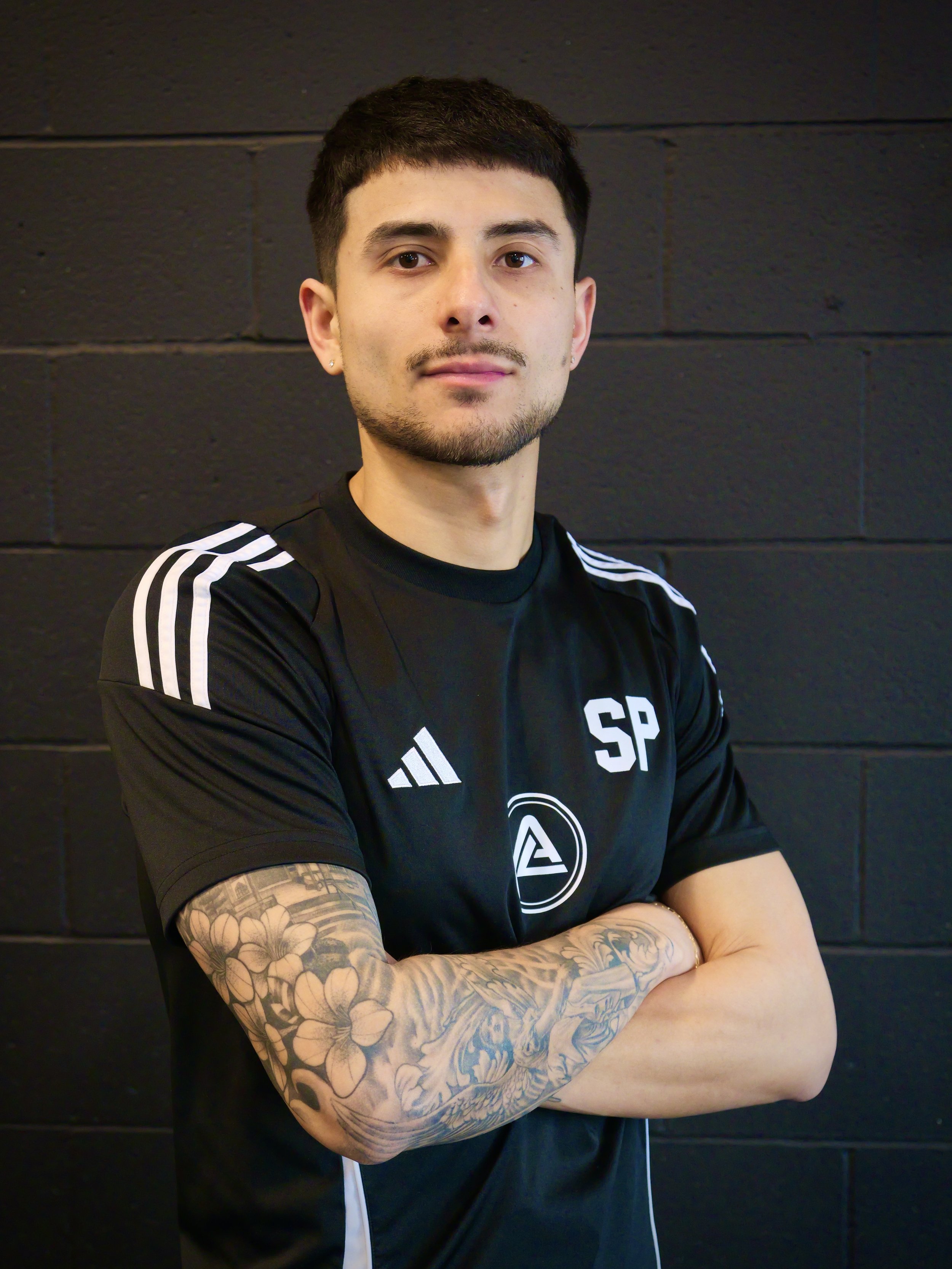 Young man with short dark hair and tattoos on his right arm, crossing his arms, wearing a black athletic shirt with white stripes on the shoulders and logo, standing against a black wall.