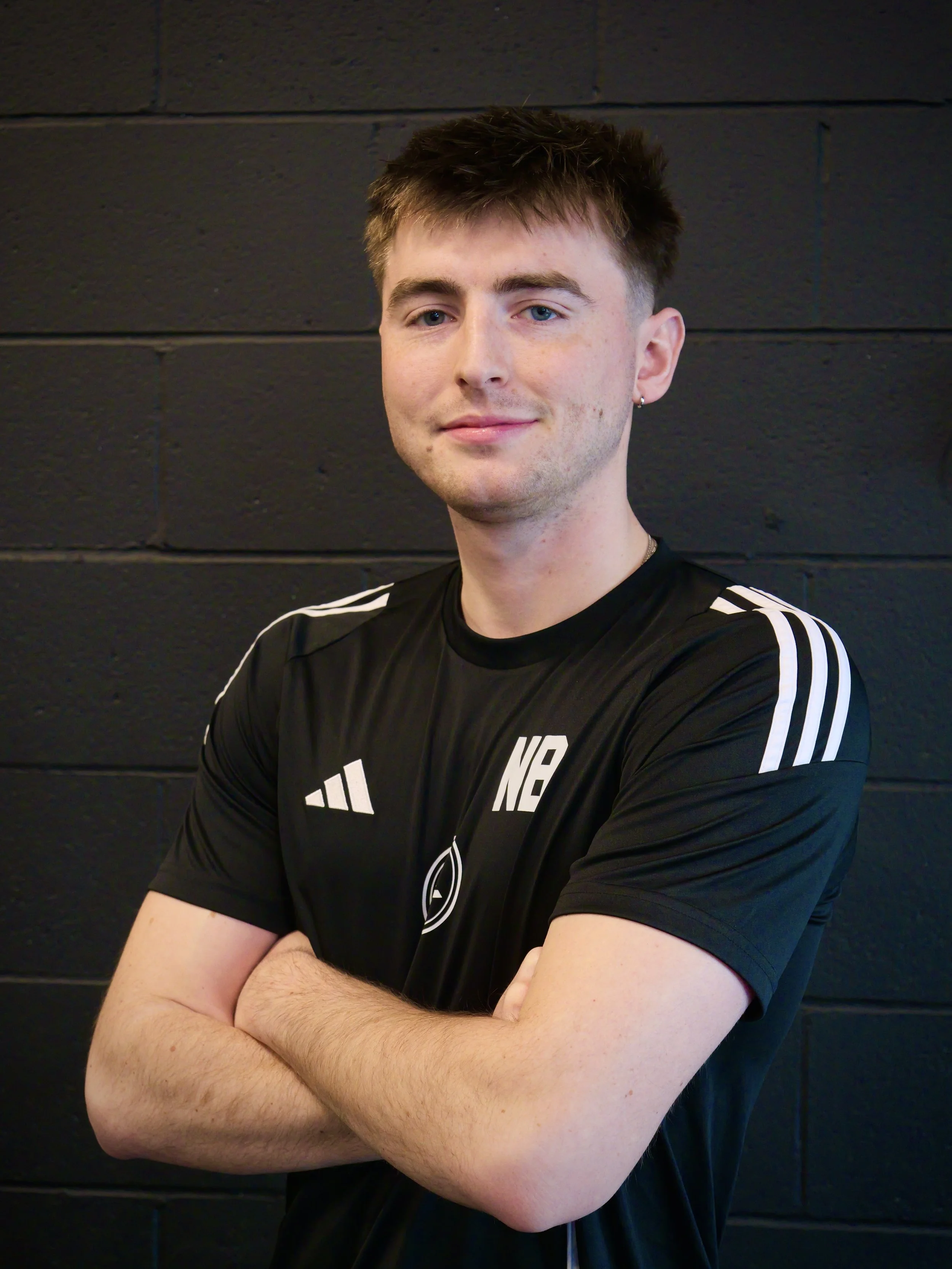 A young man with short brown hair and light skin, standing with arms crossed, wearing a black sports jersey with white accents, against a black painted brick wall.