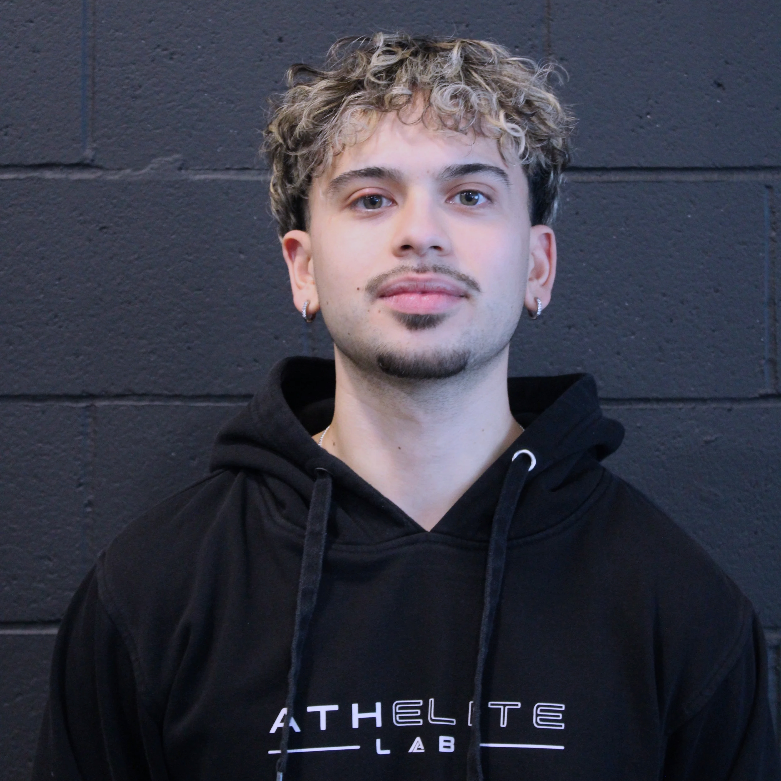 A young man with curly blonde hair, earrings, and a small goatee, wearing a black hoodie with 'Athlete Lab' written on it, standing in front of a black brick wall.