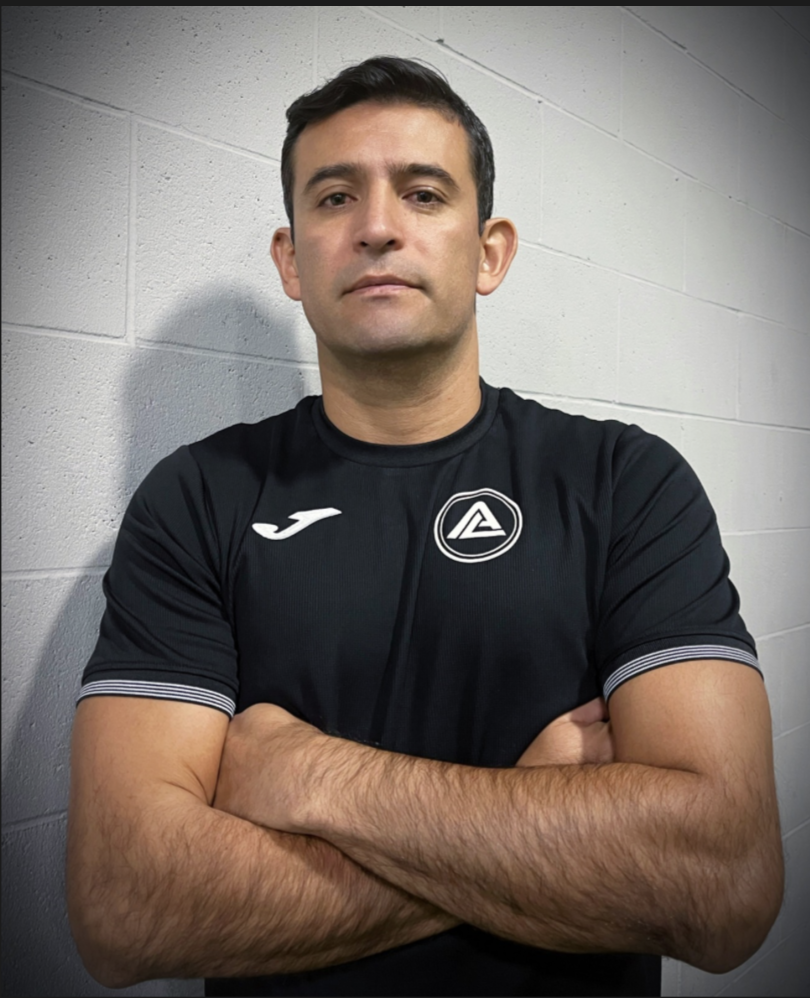Man with dark hair and light skin standing against a white brick wall, arms crossed, wearing a black sports shirt with a white logo on the chest and a white logo on the right shoulder.