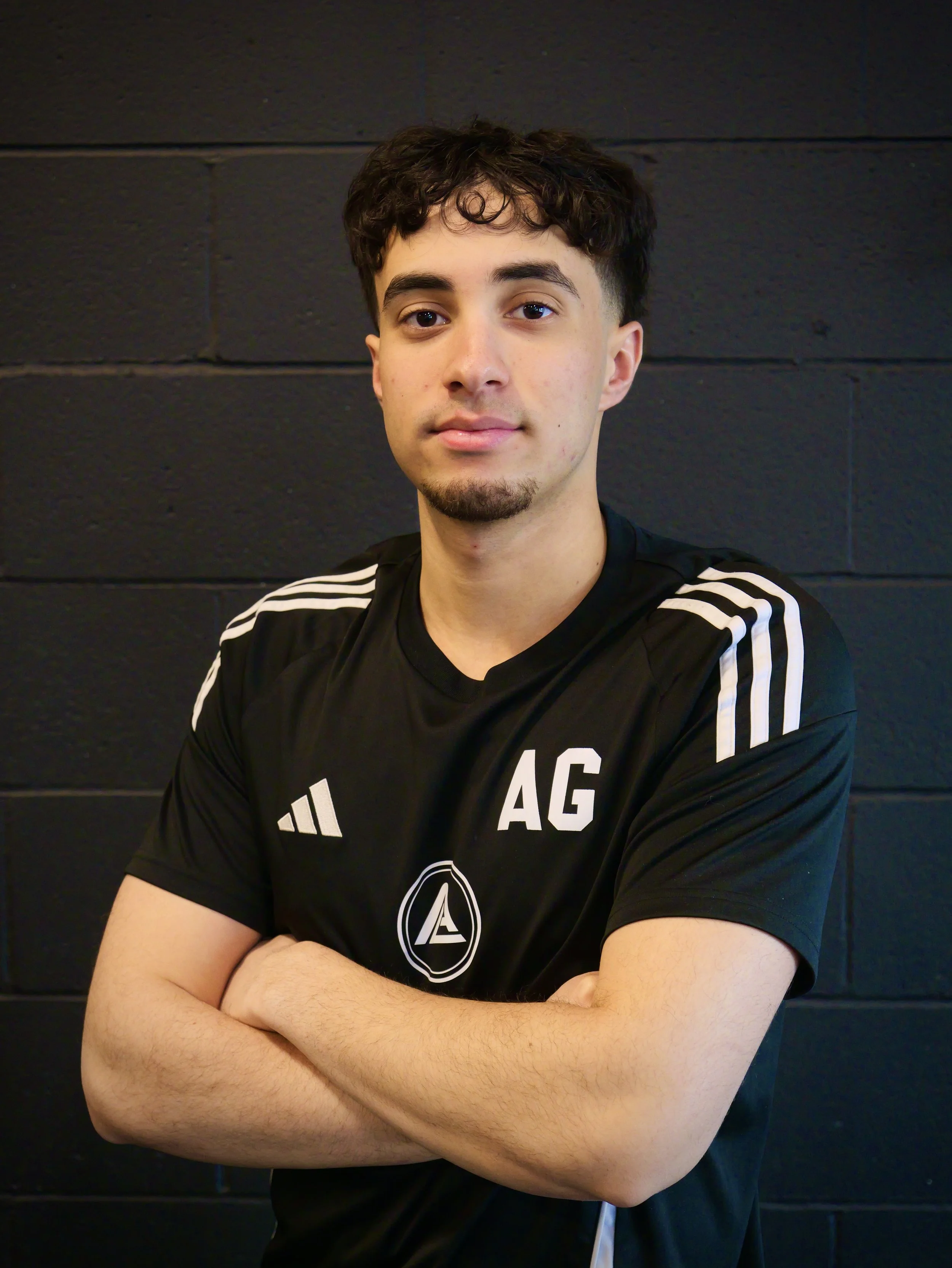 A young man with dark, curly hair and a slight beard, wearing a black athletic shirt with white stripes and the letters 'AG' on the chest, posing with arms crossed in front of a black brick wall.