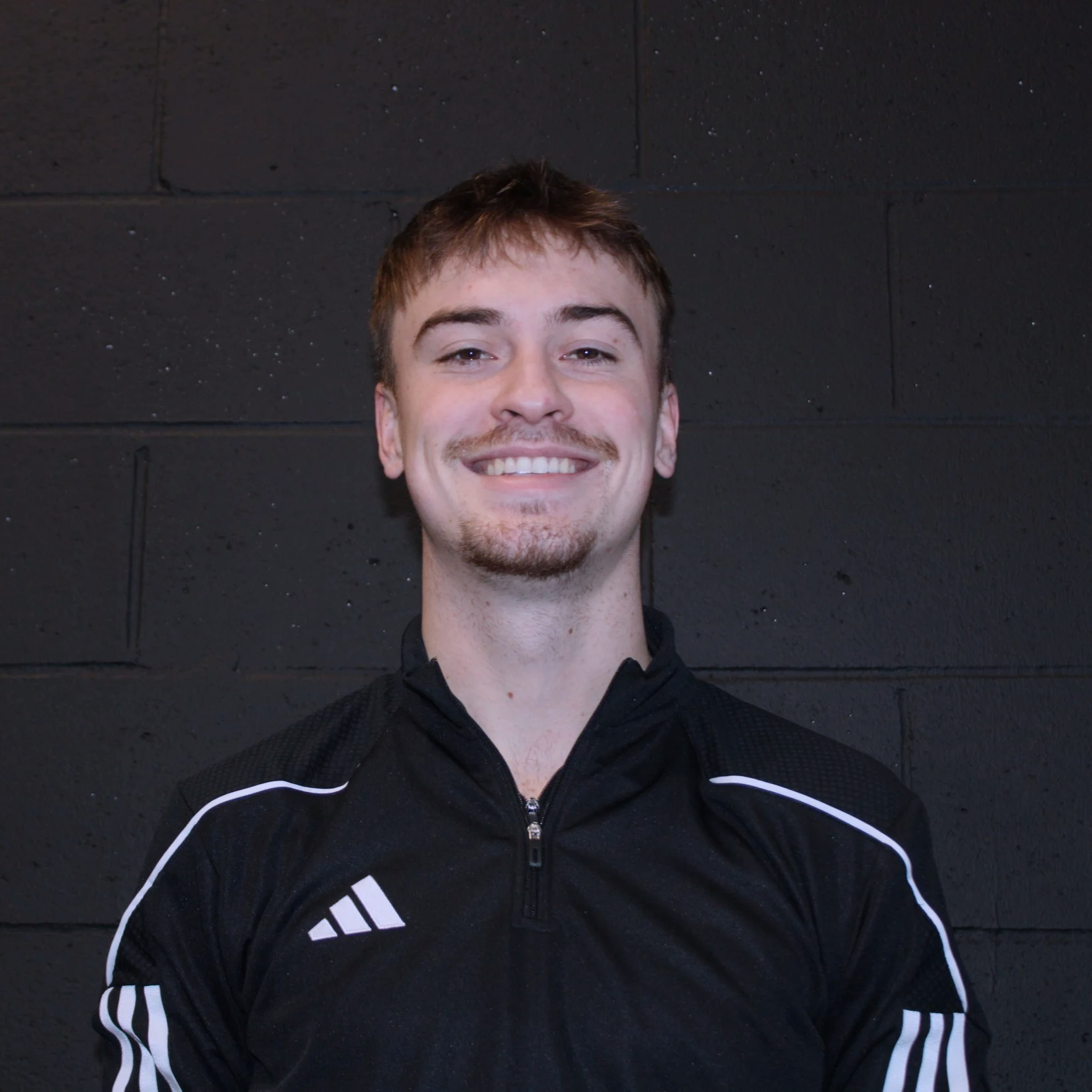 Smiling young man in black athletic jacket standing against black brick wall.