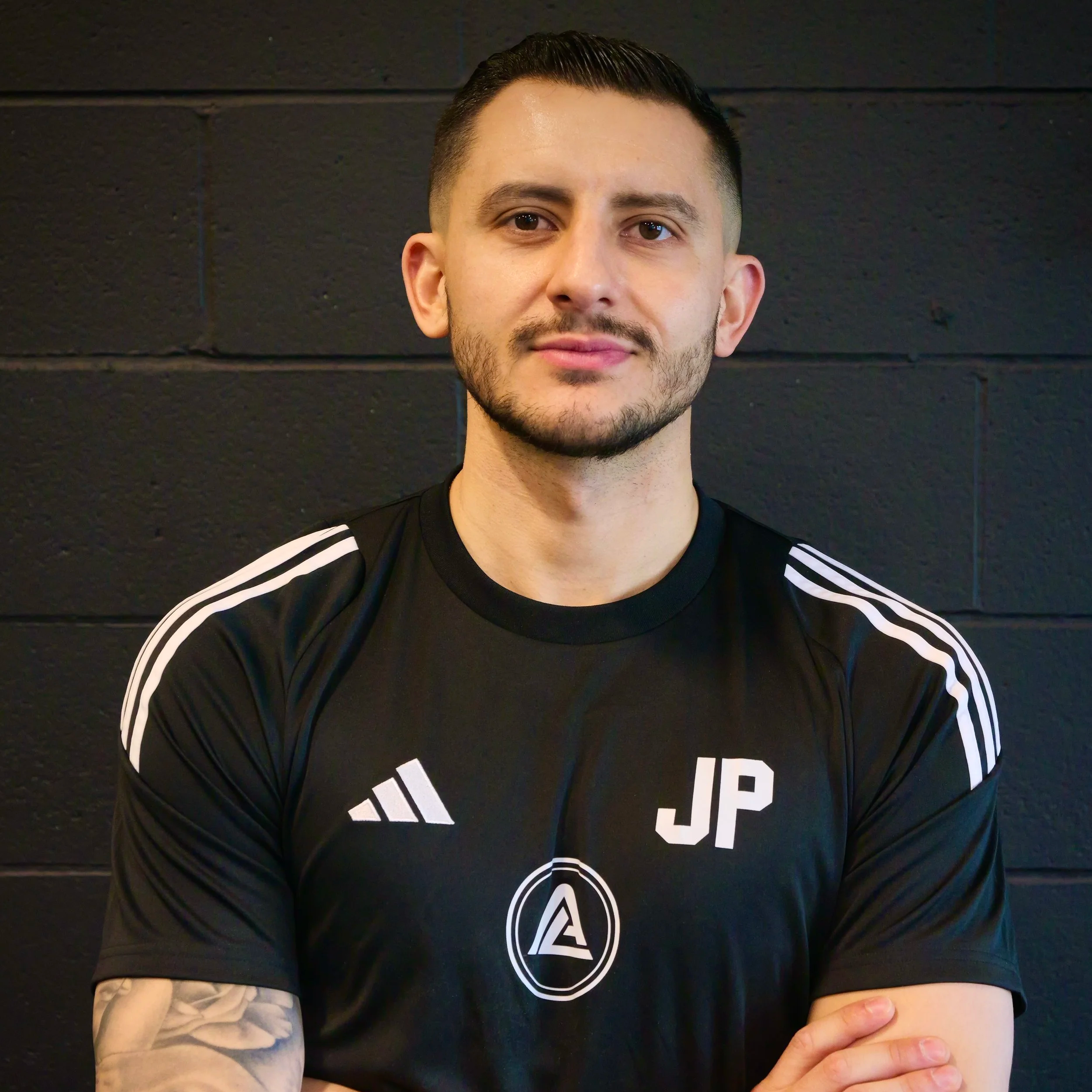 A young man with short dark hair and a beard, wearing a black sports shirt with white stripes on the shoulders and the initials 'JP' on the chest, standing in front of a black brick wall.