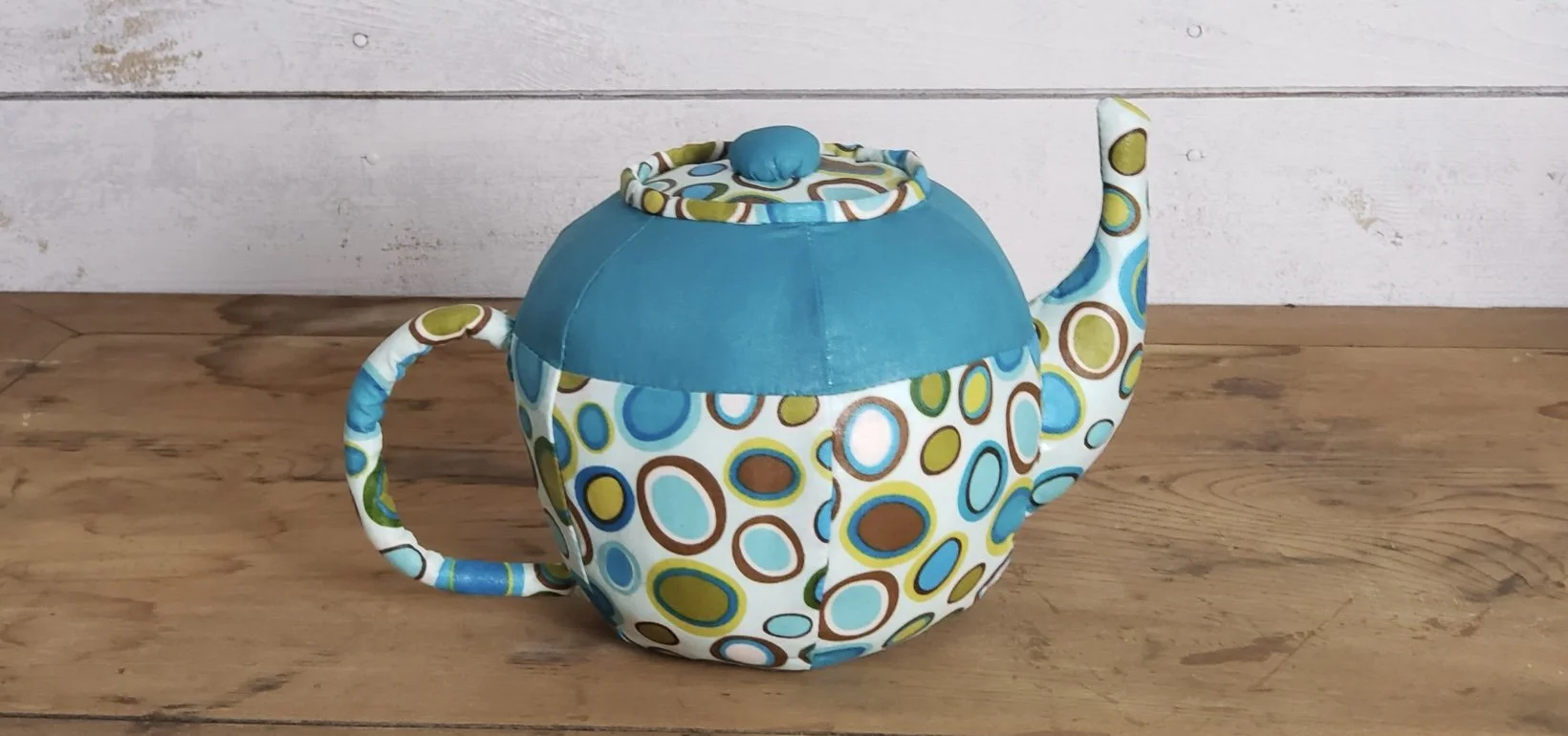 Colorful fabric teapot-shaped purse with blue, brown, and green circles, placed on a wooden surface with a white wood-paneled background.