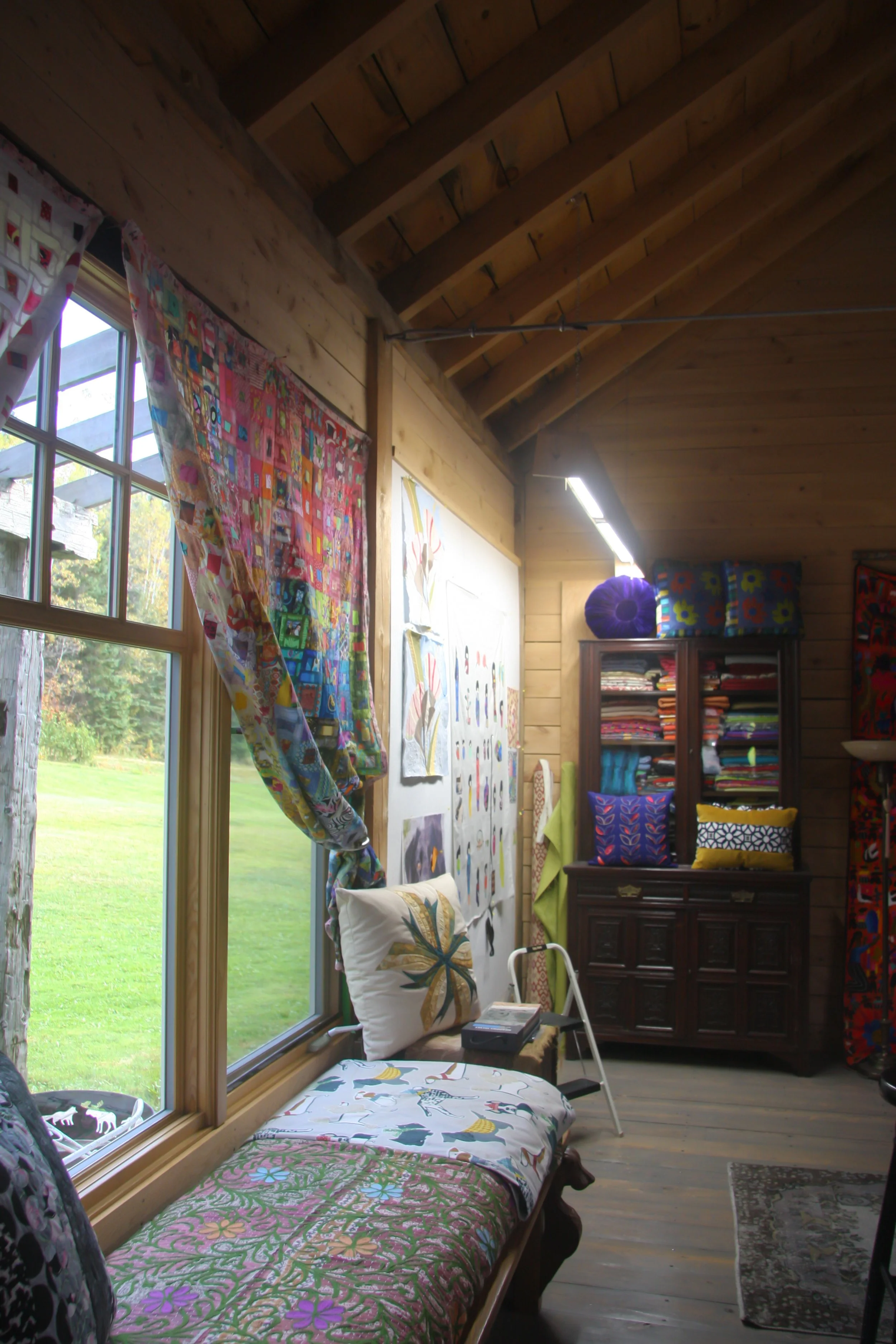 A cozy craft room with wooden walls and ceiling. It features a large window with floral curtains, a bench with colorful bedding, a white step stool, and a dark wood cabinet filled with colorful fabric and cushions. Artworks and vibrant quilts decorate the walls.