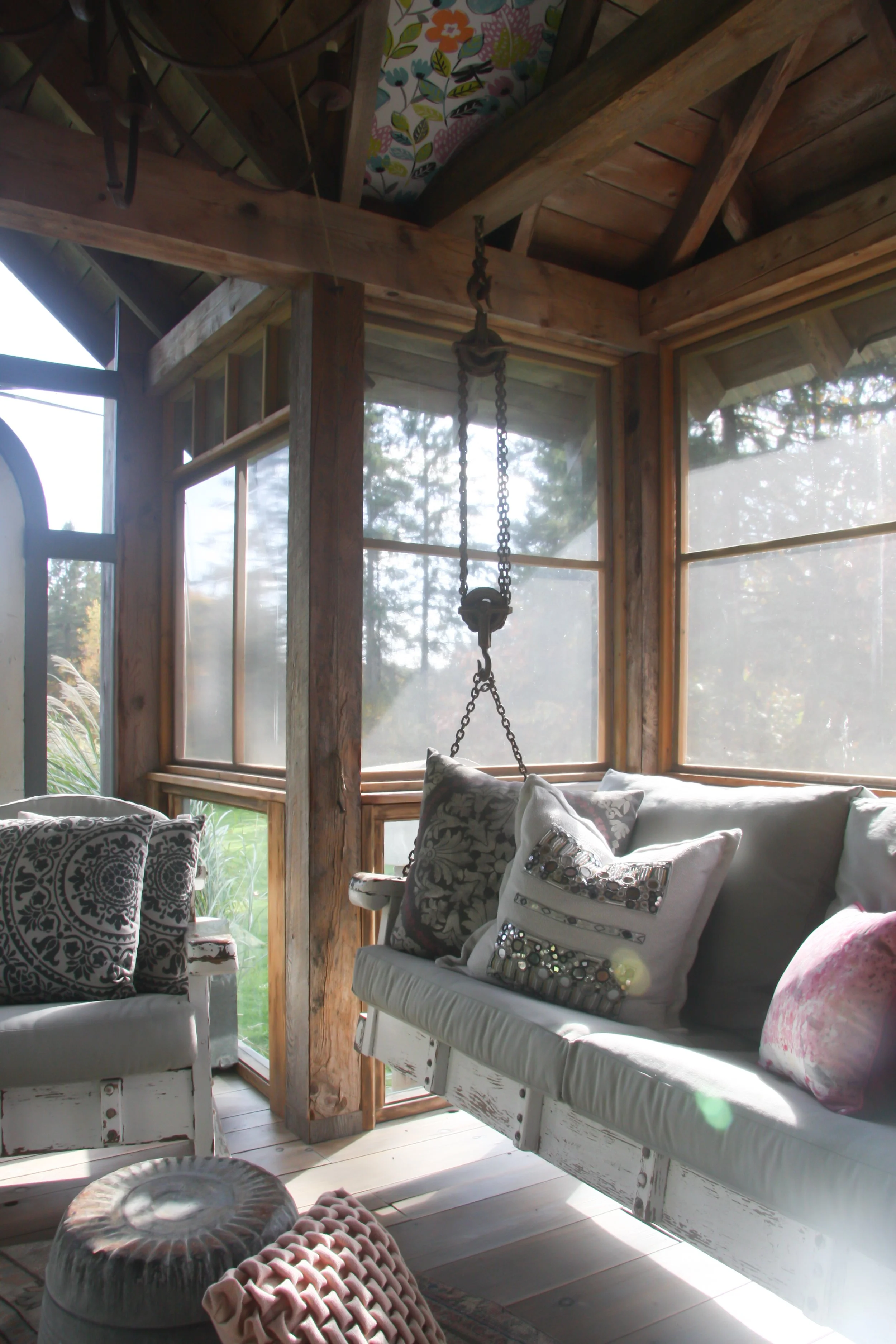 Interior of a cozy sunroom with wooden beams and large windows, decorated with patterned cushions on white-painted furniture, sunlight streaming in with visible lens flare.