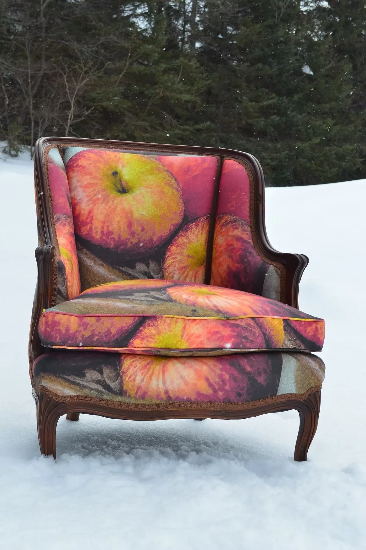 A vintage armchair with ornate wooden frame and floral fabric cushions, sitting on snow with trees in the background.