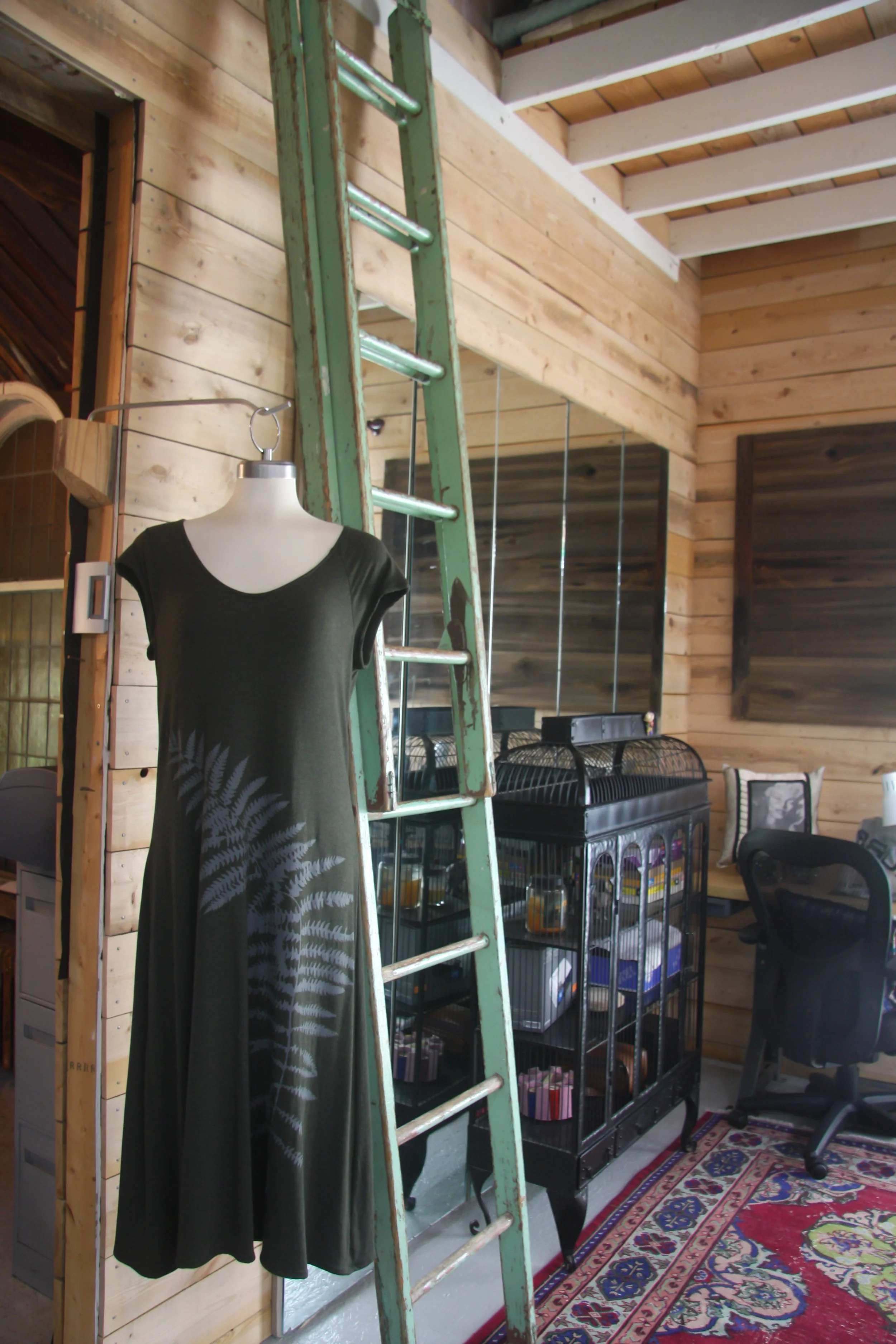 Interior of a room with wooden walls, a green ladder, a mannequin with a black dress that has a fern leaf design, a black pet cage, a black office chair, and a patterned rug.