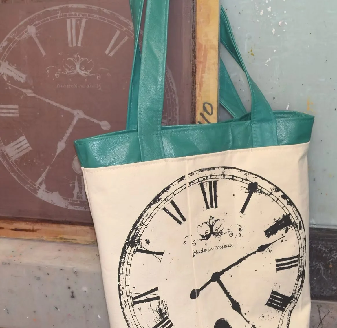 White tote bag with a black distressed clock face and Roman numerals, green handles, and a small decorative emblem that says "Made in Roseau" is hanging in front of a framed clock print.