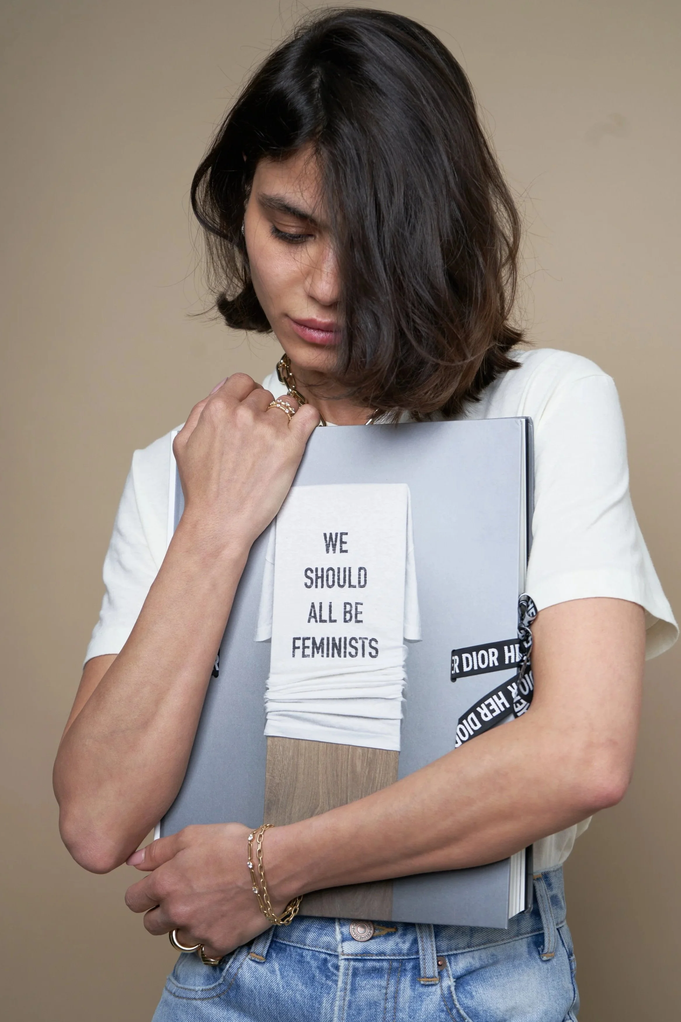 A woman with dark, shoulder-length hair holding a large book or folder against her chest, with an art piece attached to it showing a stack of white T-shirts with the message "WE SHOULD ALL BE FEMINISTS." She is wearing a white shirt, jeans, and gold 