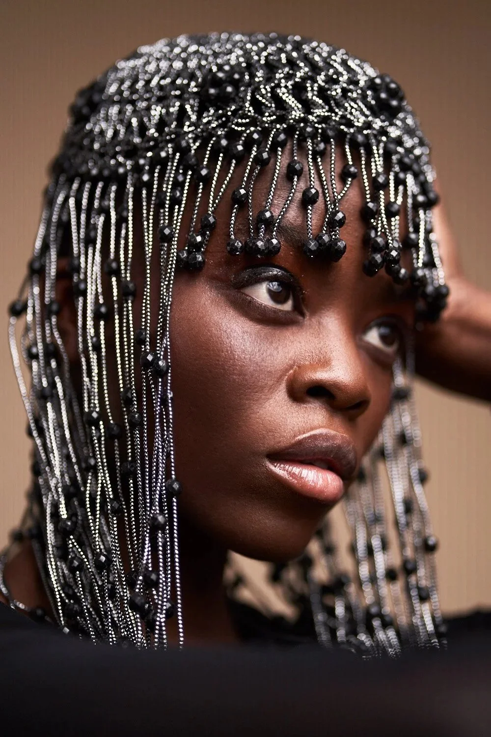 Close-up of a woman with intricate braided hairstyle adorned with black beads and silver chains, with her hand behind her head and looking slightly upwards.