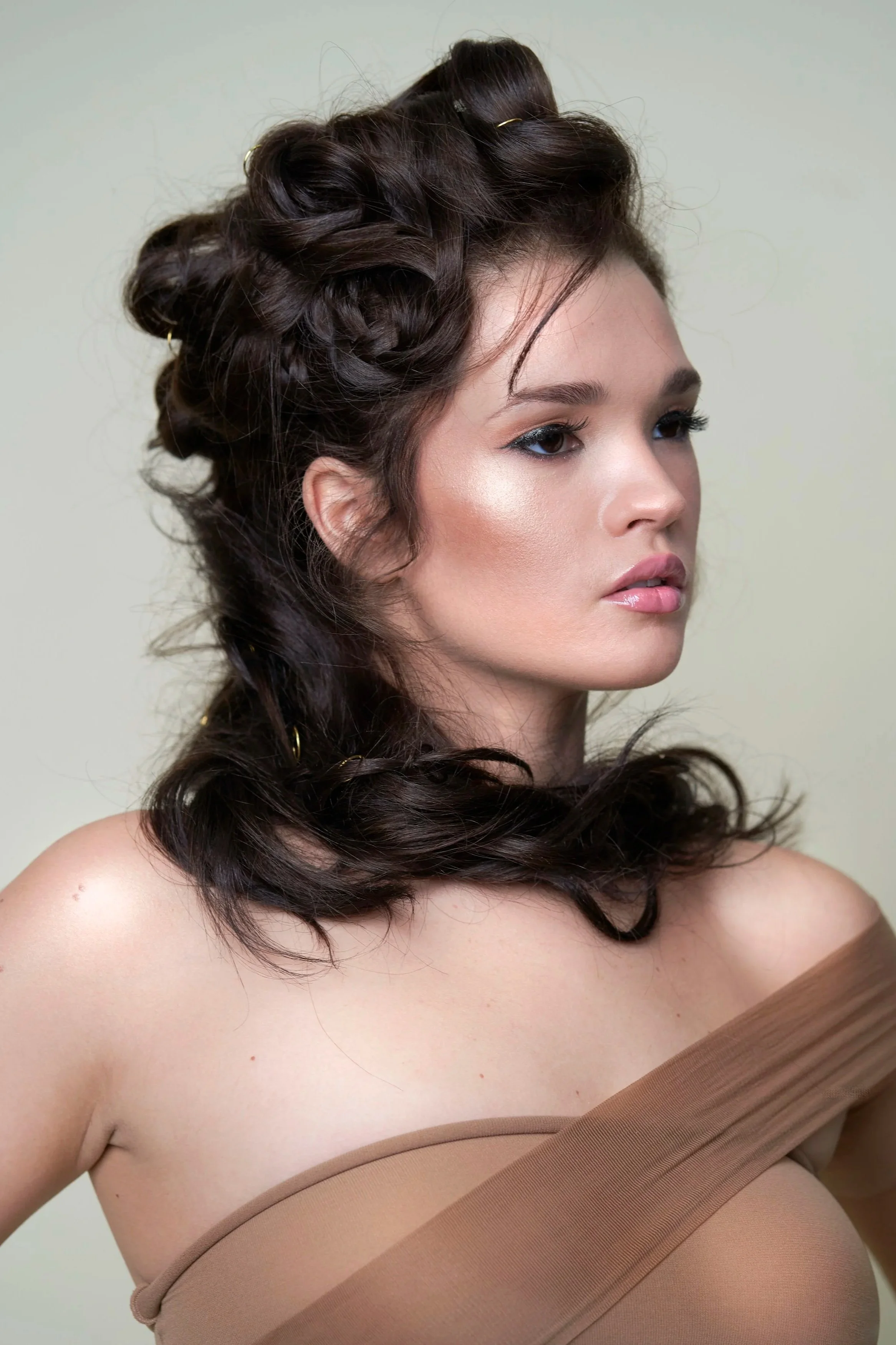 A woman with dark styled hair and subtle makeup, wearing a strapless beige top, looking to the side against a plain background.