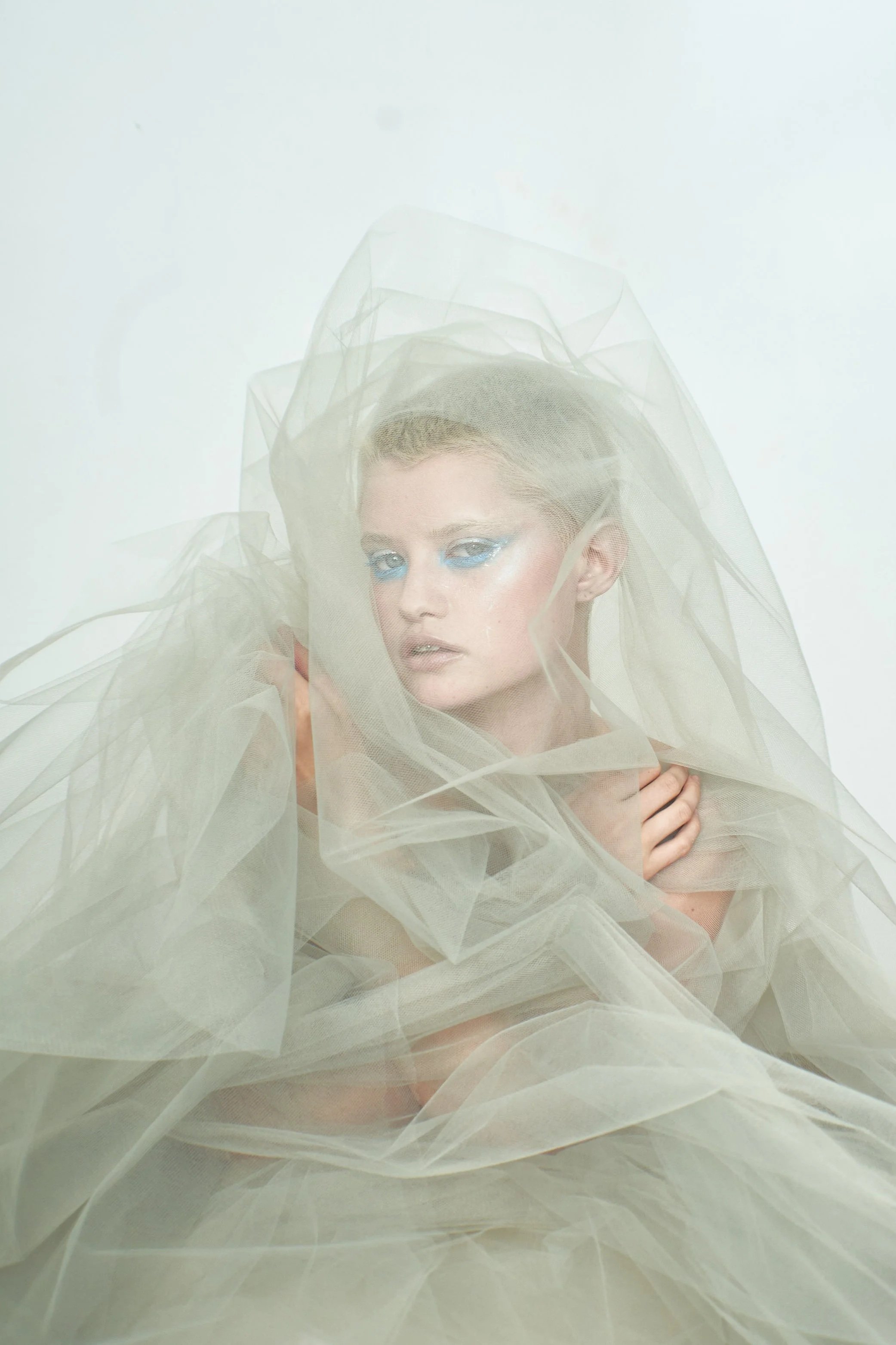 A woman with blue eye makeup and blonde hair, wrapped in sheer, light-colored fabric against a plain white background.