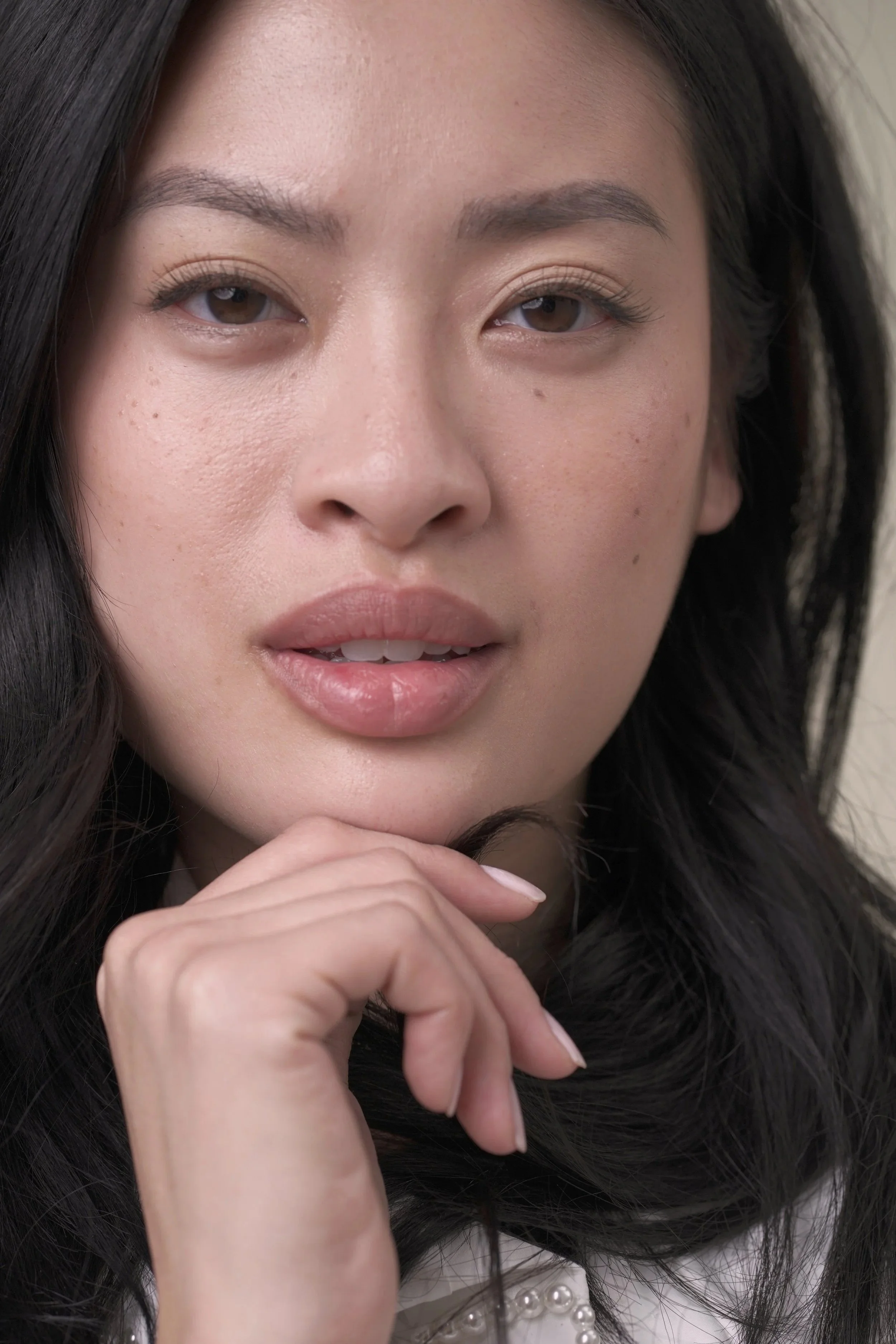 Close-up of a woman with long black hair, light skin, and brown eyes, looking directly at the camera with her hand gently touching her chin.