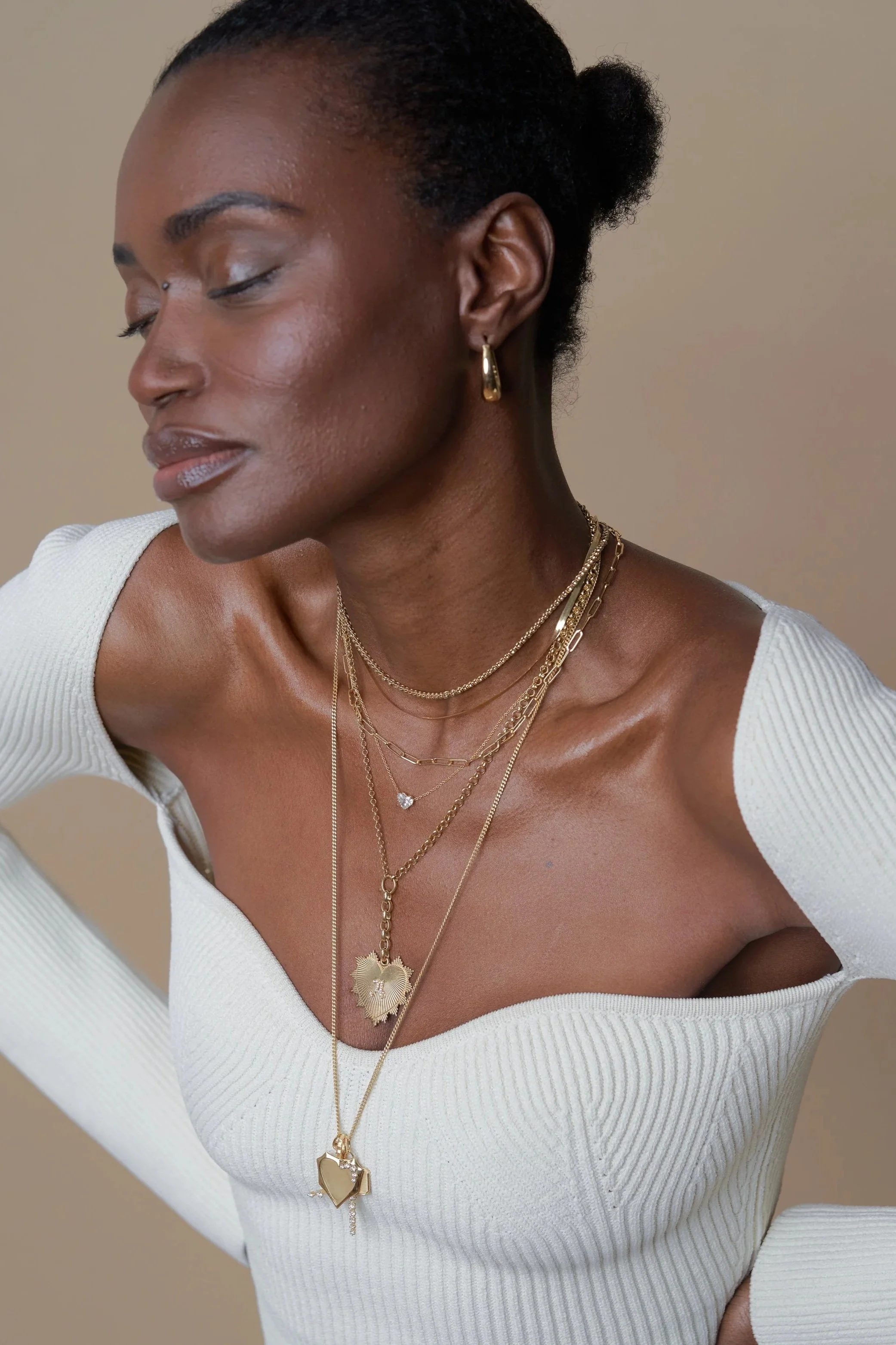 A woman with dark skin and short black hair styled in a small bun, wearing multiple layered gold necklaces, gold earrings, and a white off-the-shoulder sweater.