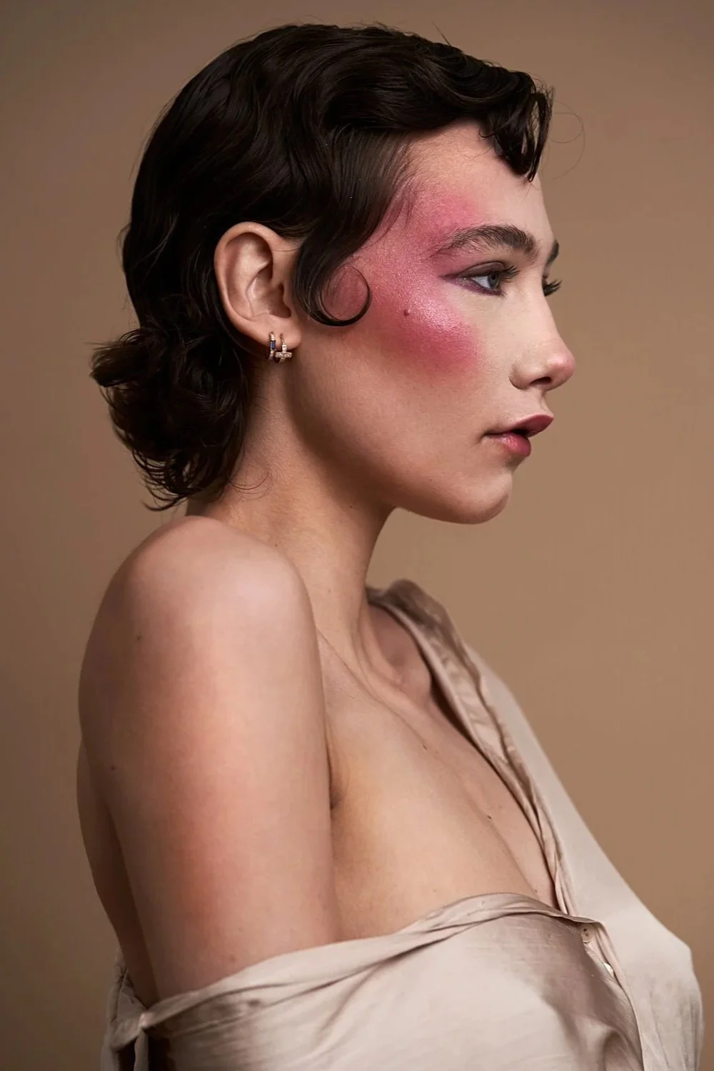 A woman with short dark hair and earrings, wearing a beige off-shoulder top, has a visible red rash or irritation on her cheek and side of her face.