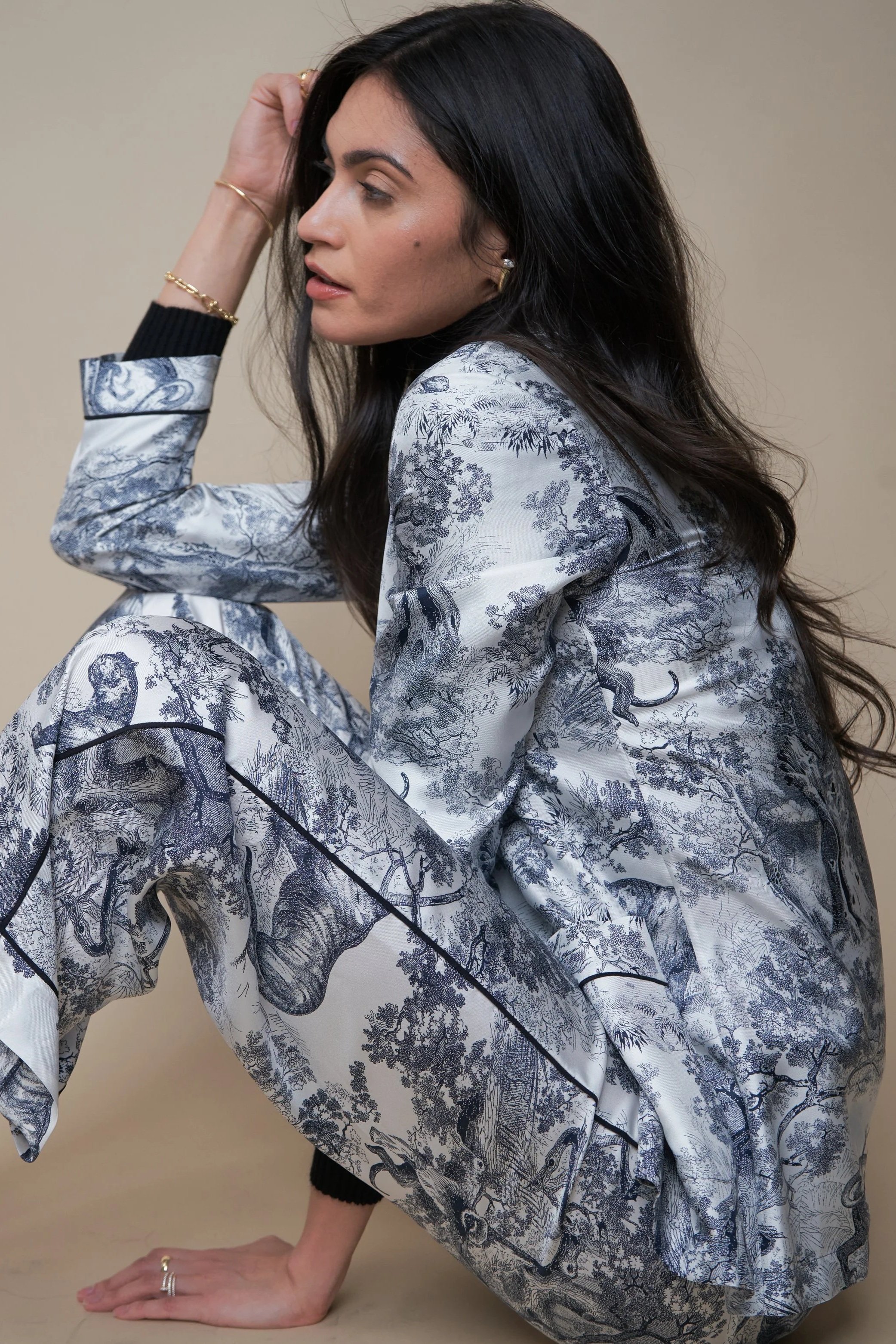 A woman with long dark hair is crouching with one hand on the floor and the other touching her head. She is wearing a matching blazer and pants with a detailed black and white forest and animal print.