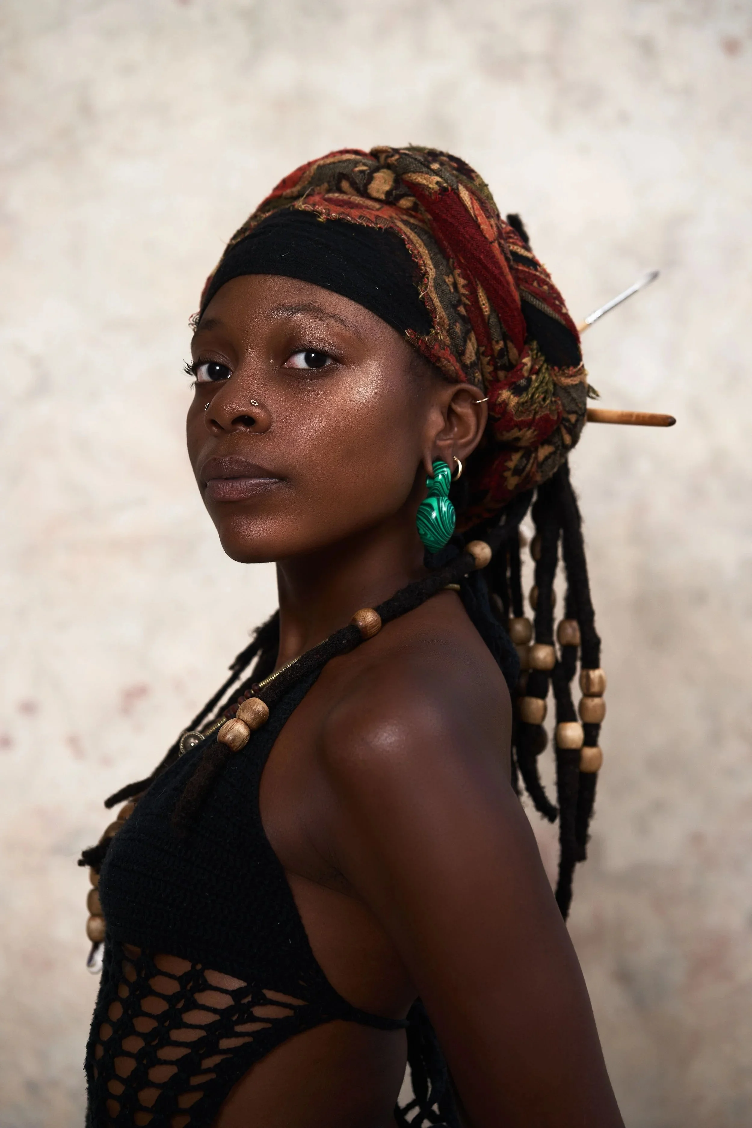 A woman with dark skin and dreadlocks wearing a headscarf, green earrings, and a black crocheted top, looking over her shoulder against a neutral background.