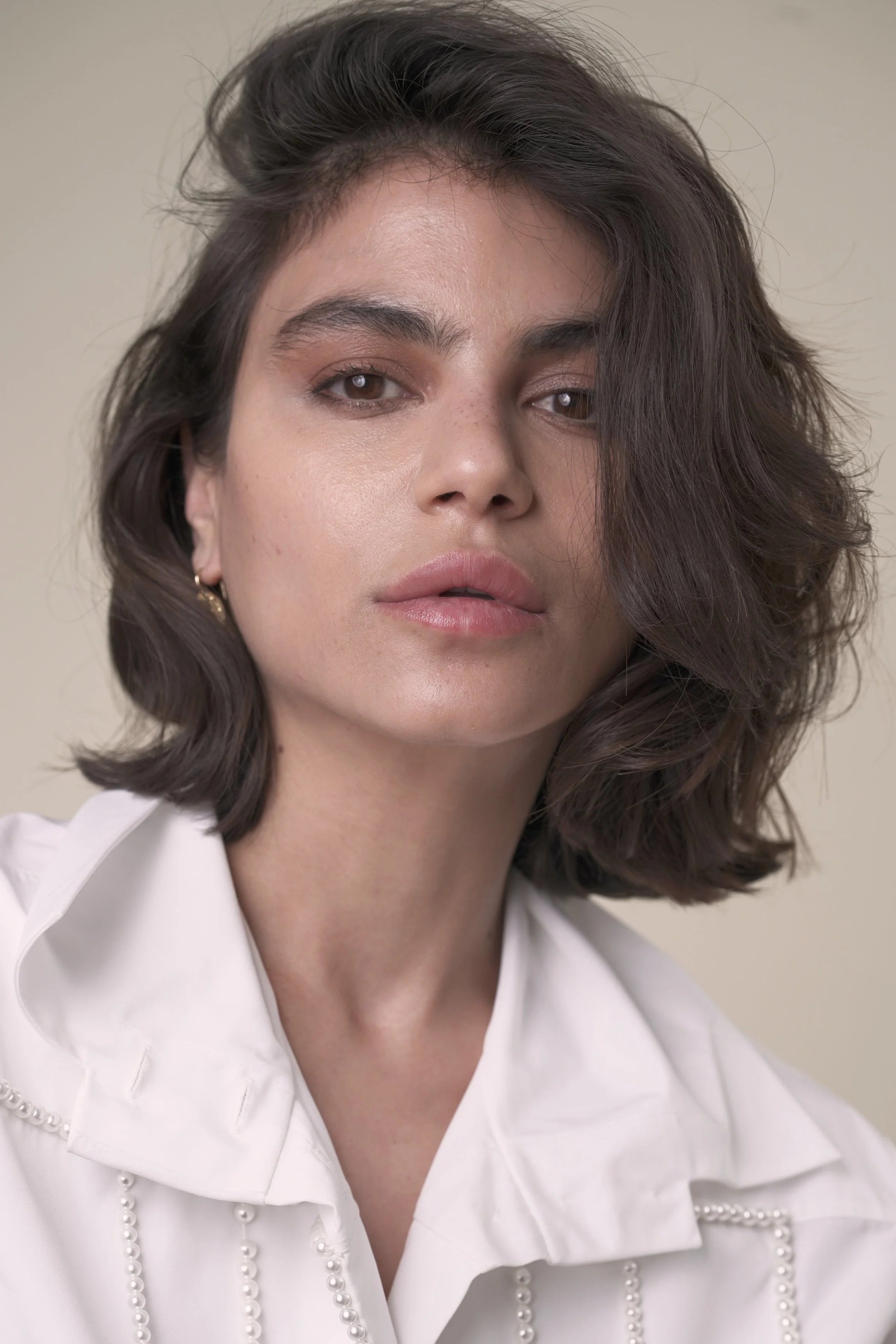 A woman with shoulder-length dark brown hair, light skin, and wearing a white shirt with pearl embellishments, looking directly at the camera with a neutral expression.