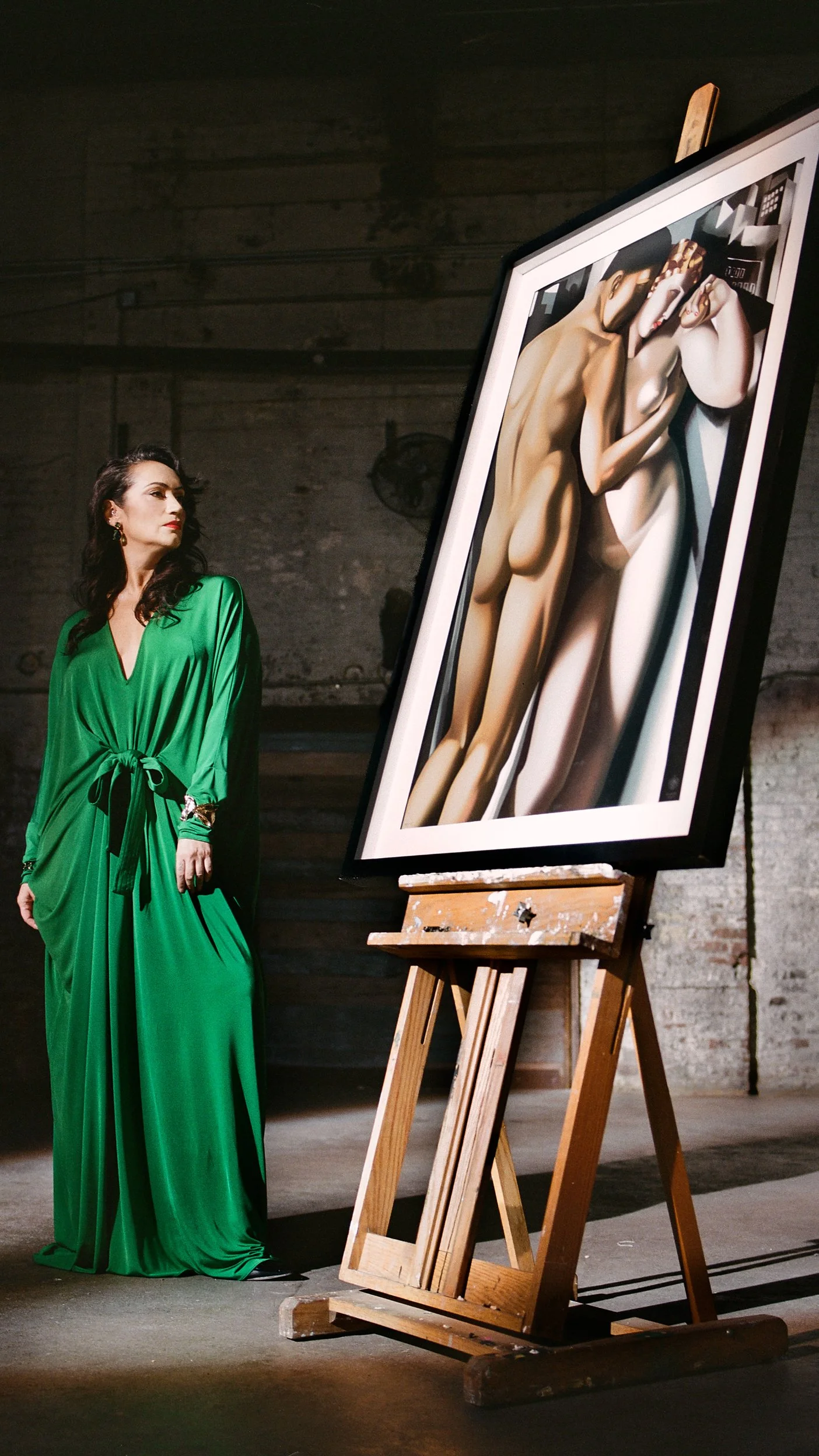 A woman in a green dress observing a large painting of abstract nude figures on an easel in an art studio.