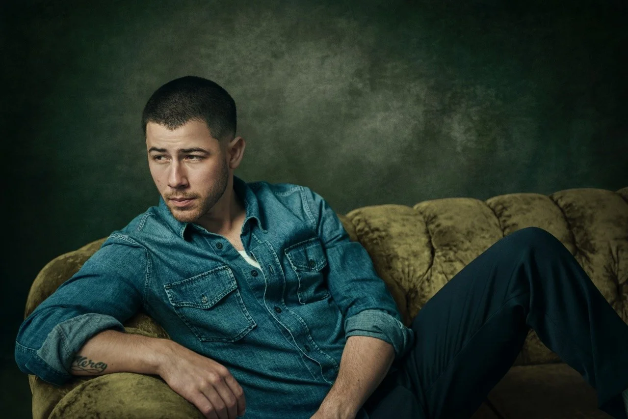 A young man with short dark hair and a tattoo on his left forearm sits on a vintage green velvet couch, wearing a denim shirt and black pants, looking to his left with a neutral expression, against a textured dark green background.