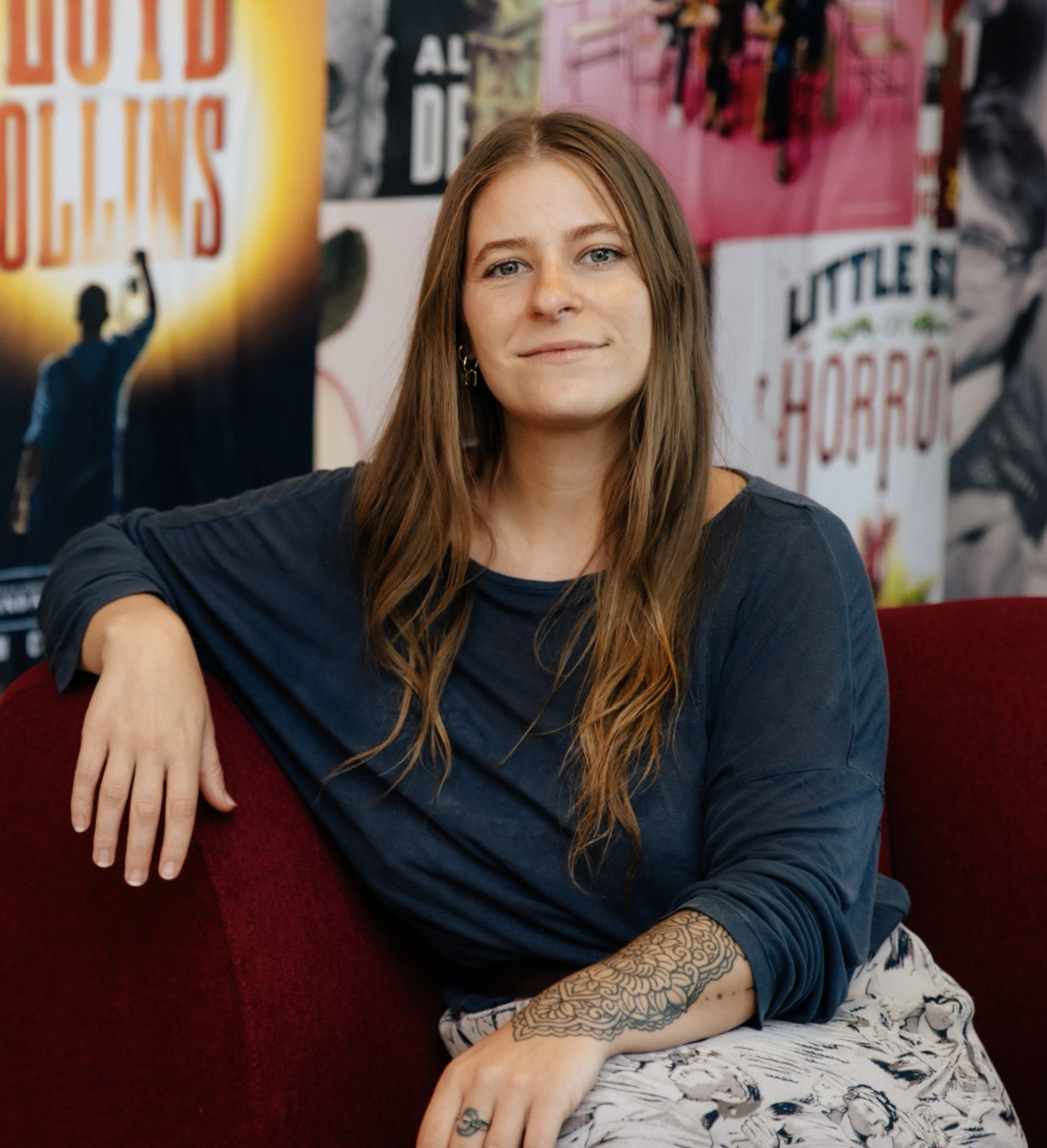 A young woman with long brown hair and light skin sitting on a red couch in front of a collage of colorful posters.
