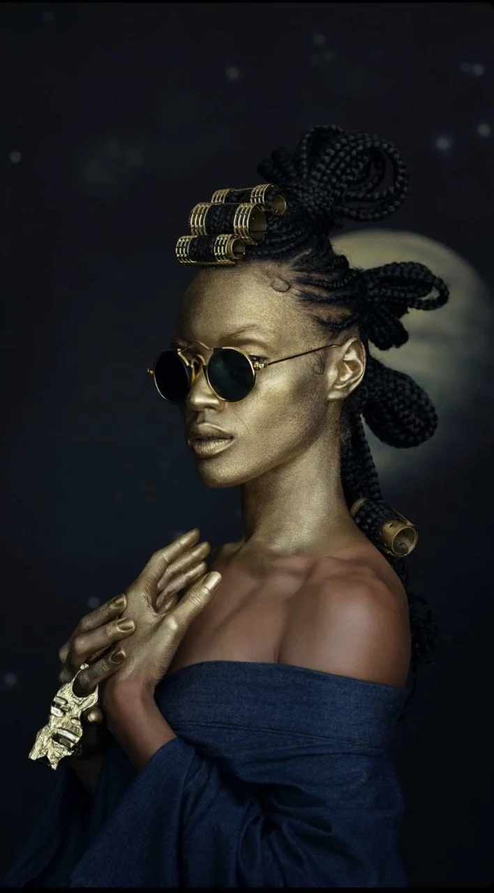 A woman with gold skin and elaborate hairstyle wearing round sunglasses, a dark off-shoulder top, and a large gold bracelet, posing with her hands crossed over her chest against a dark background with small light spots.