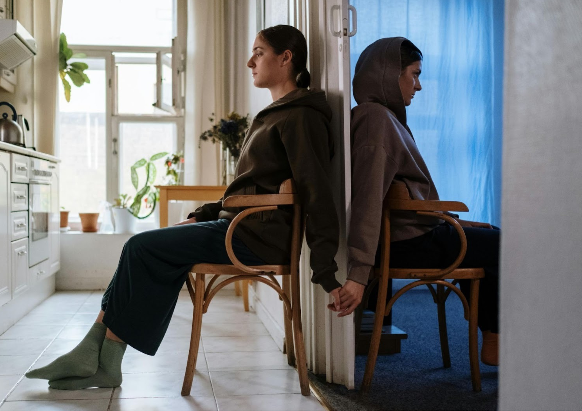 Two partners sitting back-to-back in separate rooms, quietly reaching for each other’s hands, symbolizing emotional disconnection and longing often felt in anxious attachment.
