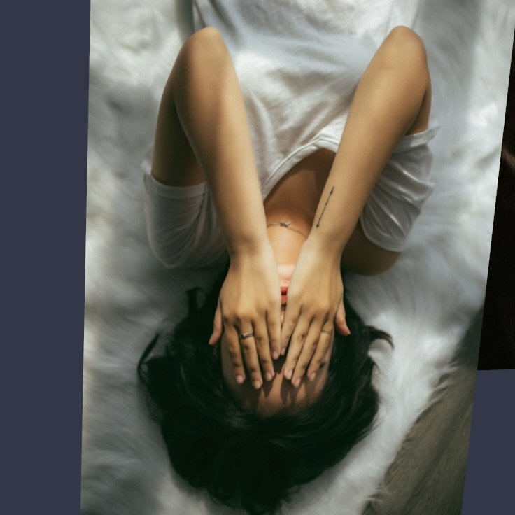 A woman in distress lying down on a white fur rug covering her face with both hands as if to hide from the world and her own emotions.