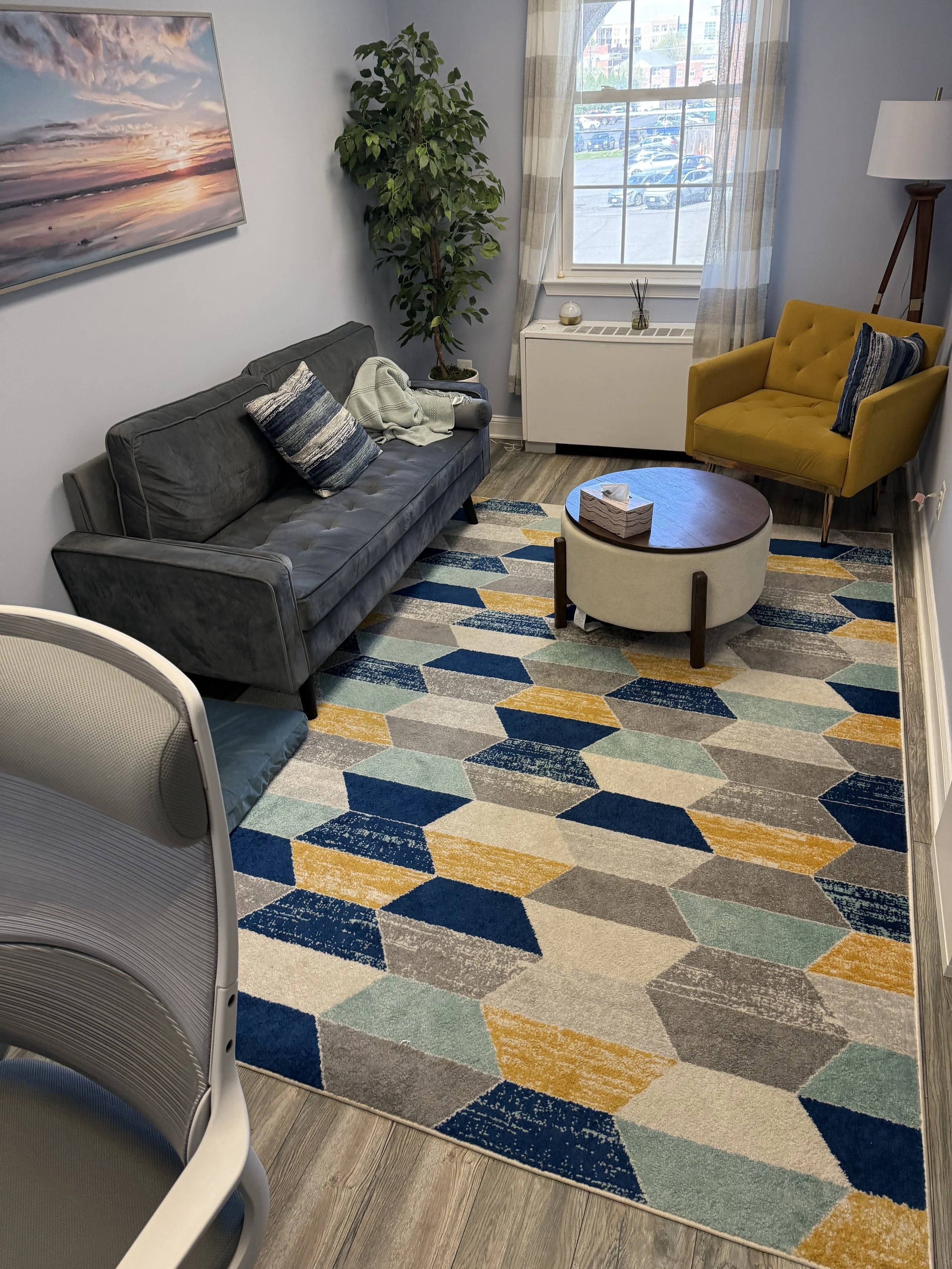 A cozy living room with a colorful geometric rug, a dark gray sofa with pillows and a throw blanket, a yellow armchair with a pillow, a round coffee table with tissue box, a tall lamp, a window with curtains, and a large painting of a sunset on the wall.